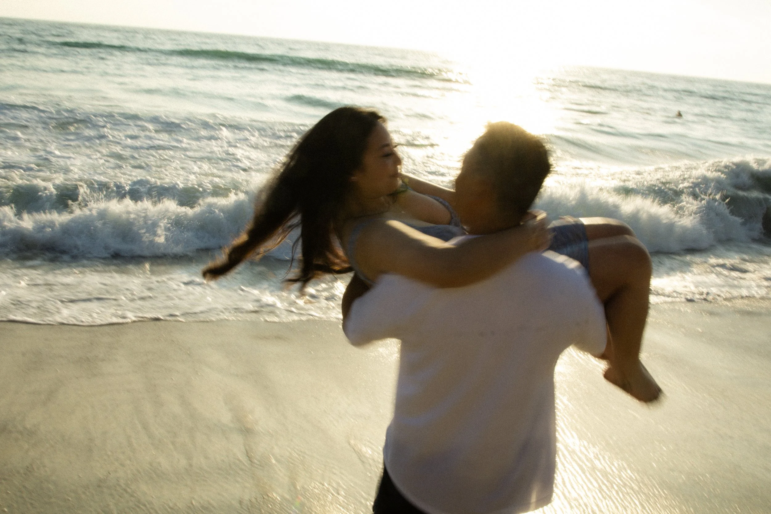 A person is carrying a woman on the beach near the water with waves, during sunset.