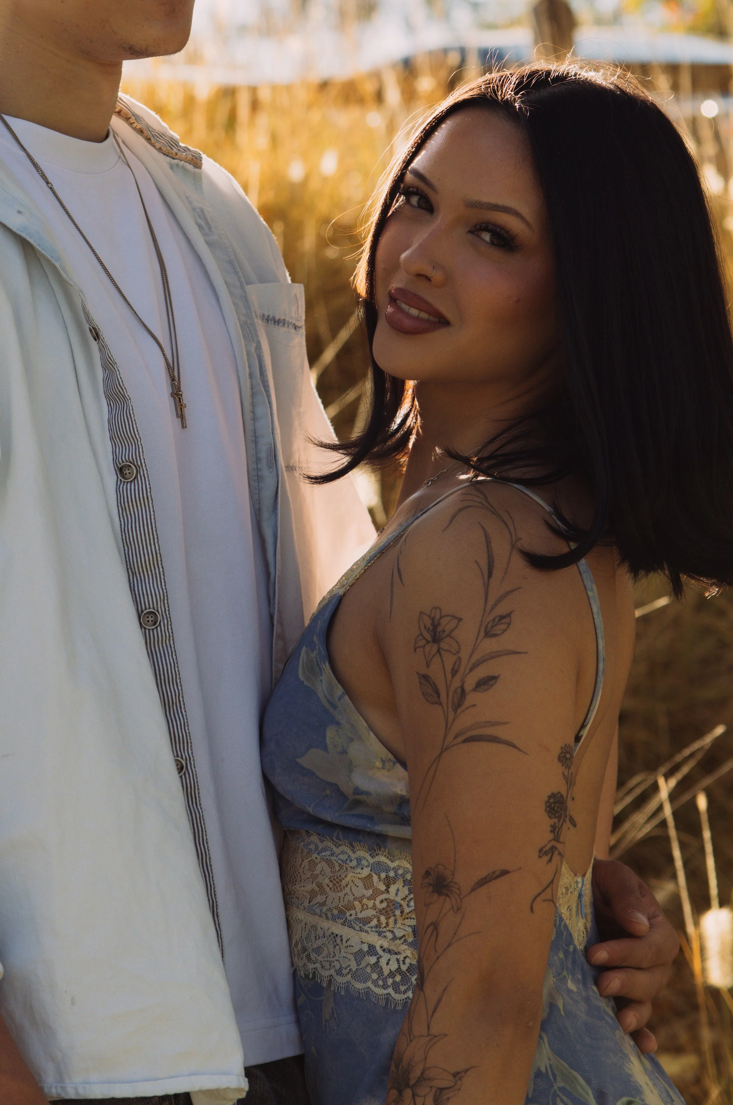 A woman with dark, shoulder-length hair and tattoos on her arm, looking at the camera, standing next to a man with a white shirt and gold chain, outdoors in a field with tall, golden grass during sunset.