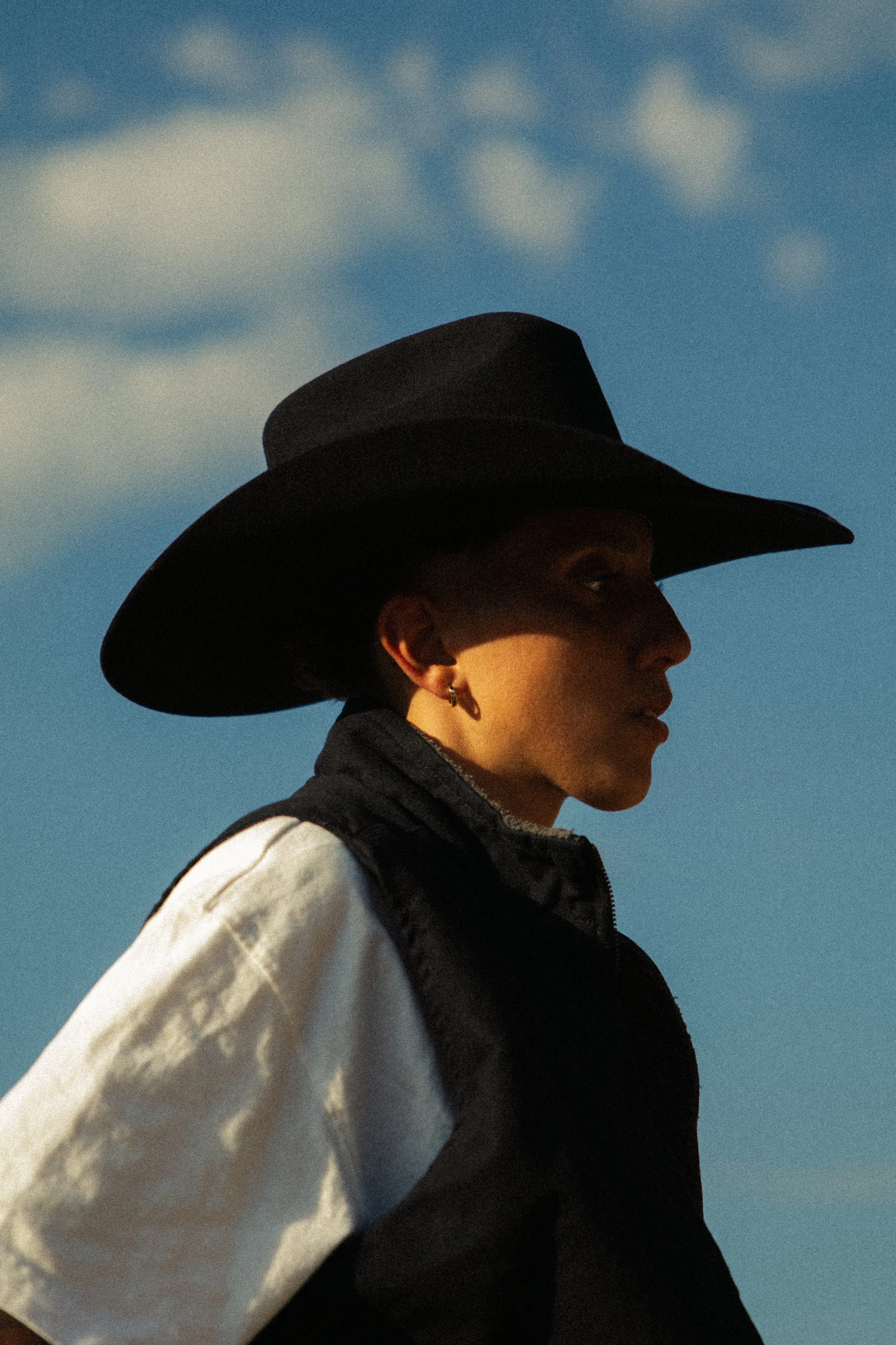 Profile of a person wearing a black wide-brimmed hat, a black vest, and a white long-sleeve shirt with black accents, standing outdoors against a blue sky with some clouds.