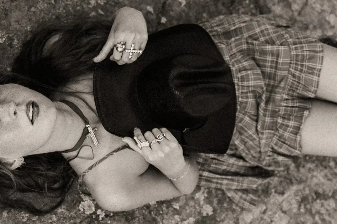 A woman lying on the ground with her eyes closed, wearing a plaid skirt, black top, and various jewelry including rings, bracelets, and a choker necklace with a cross pendant, holding a black hat.