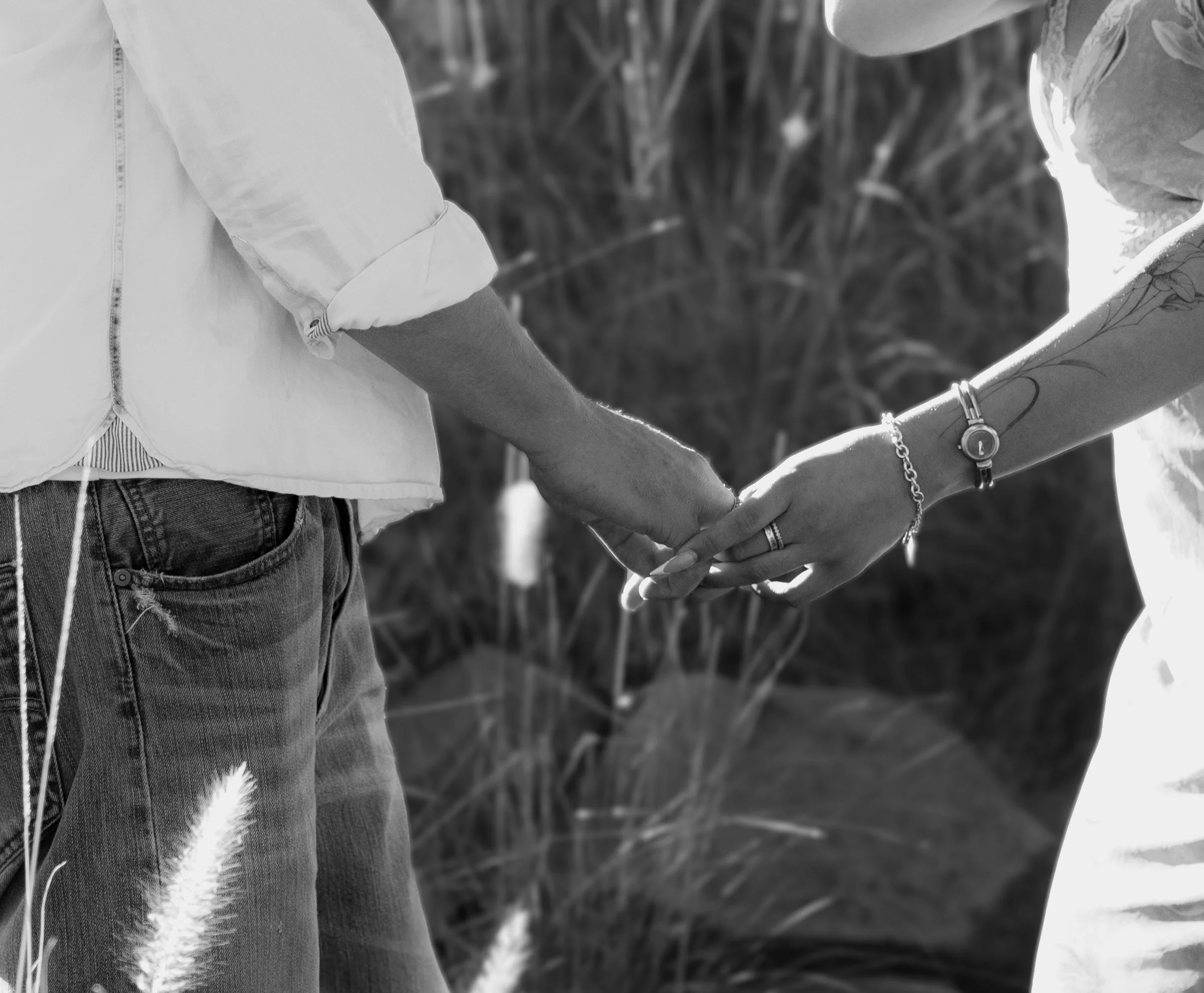 A person with a tattoo on their arm, wearing a watch and bracelet, holding hands with another person, with trees or bushes in the background.