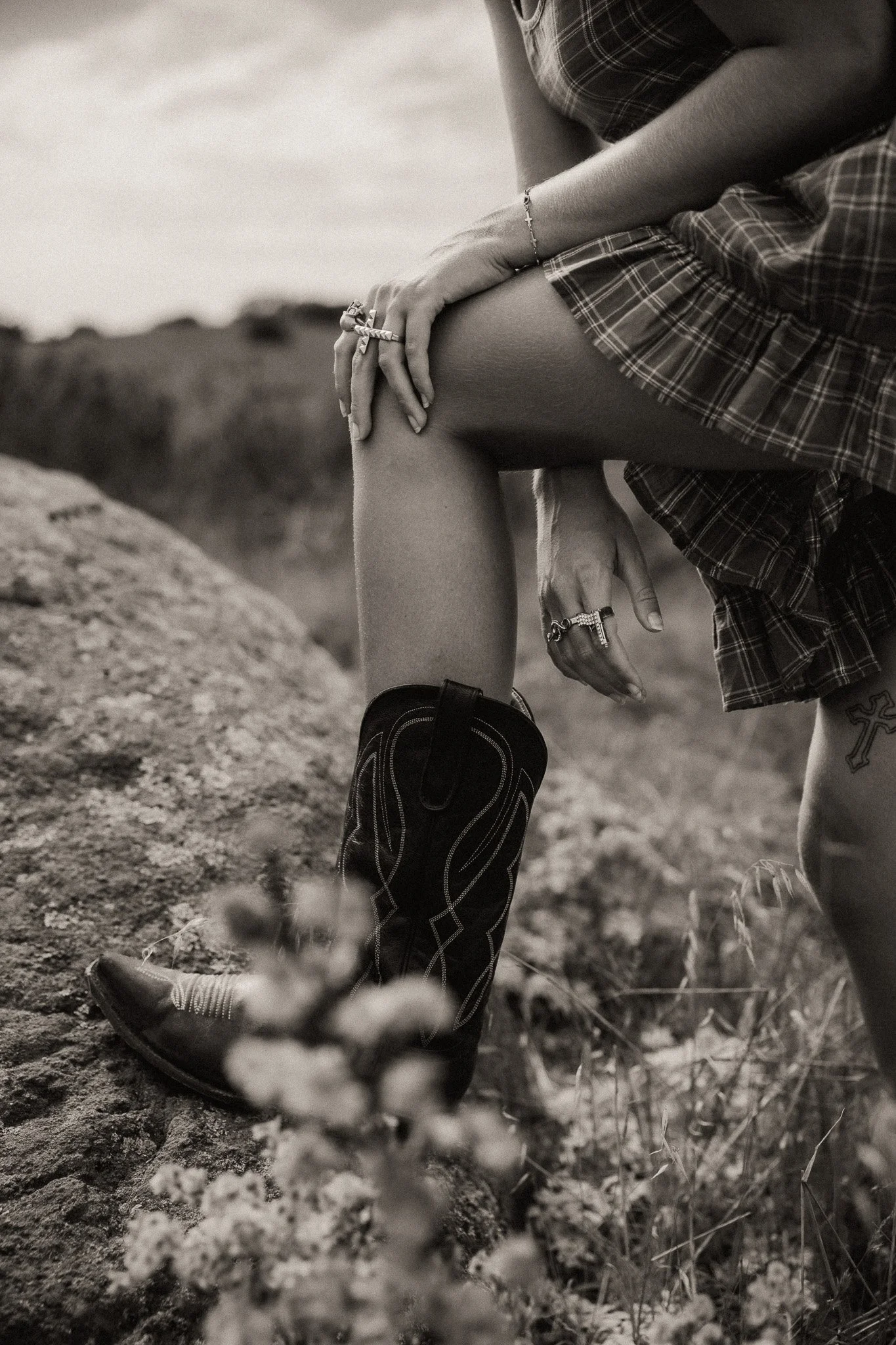 A person wearing cowboy boots, a plaid skirt, and jewelry, likely outdoors on a dirt or rocky surface in a natural setting.