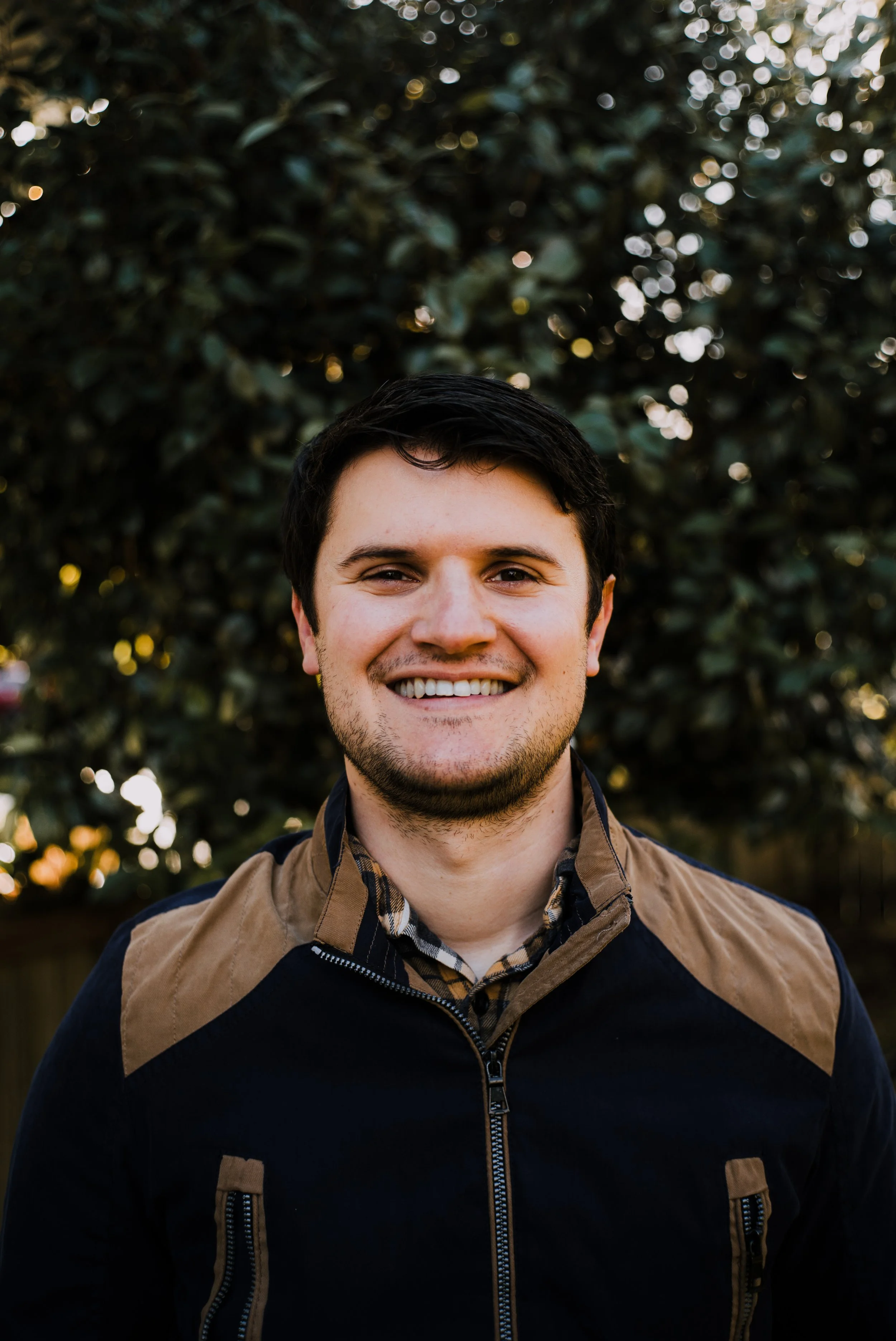A young man smiling outdoors with a blurred leafy background.