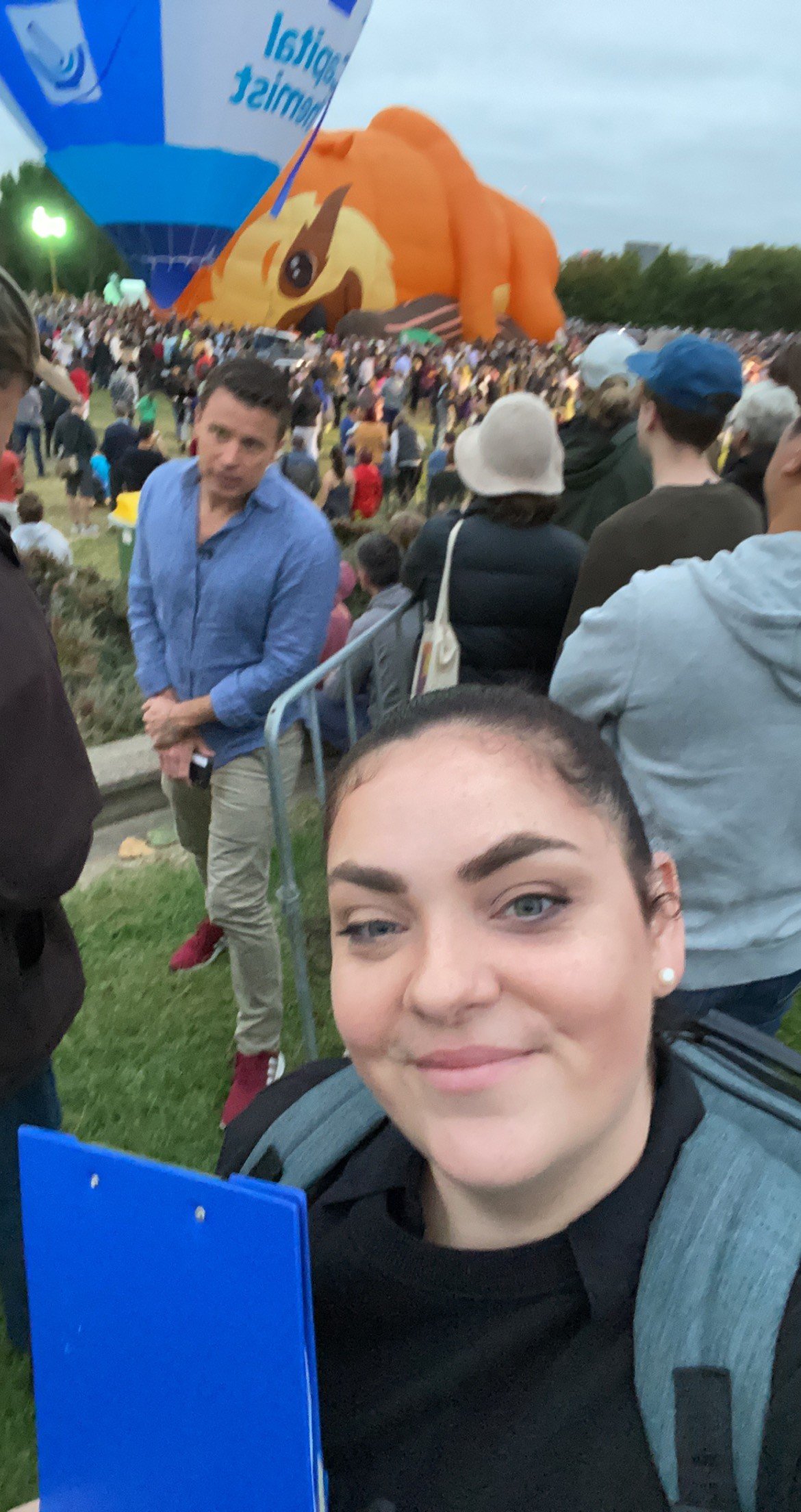 Selfie of a young woman with dark hair and blue eyes at a crowded outdoor event with a large hot air balloon shaped like a puppy in the background.