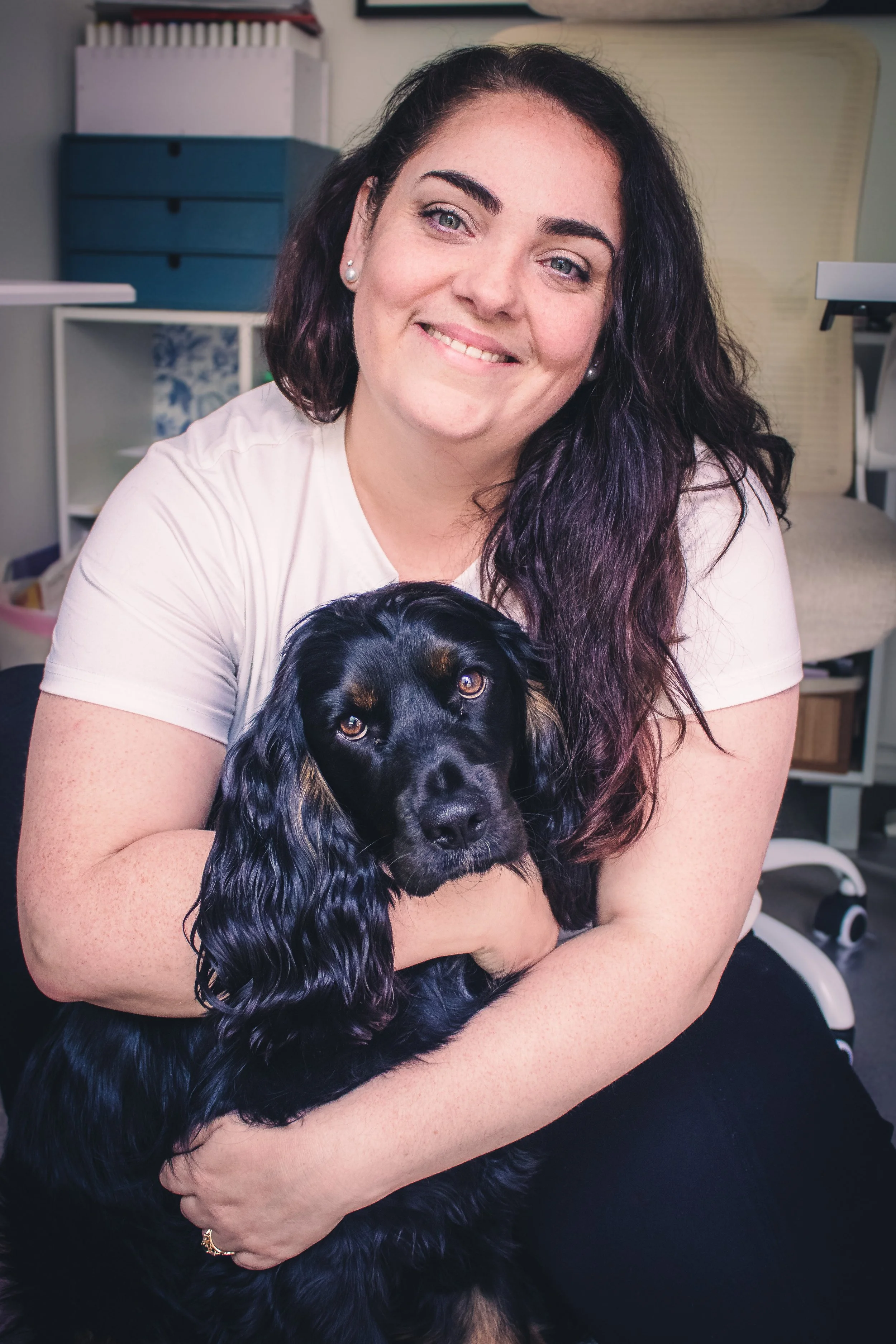 A woman with dark, wavy hair and pearl earrings smiling and holding a black and tan Gordon Setter dog with long, wavy ears. The background has a blue dresser, white shelves, and a bed.