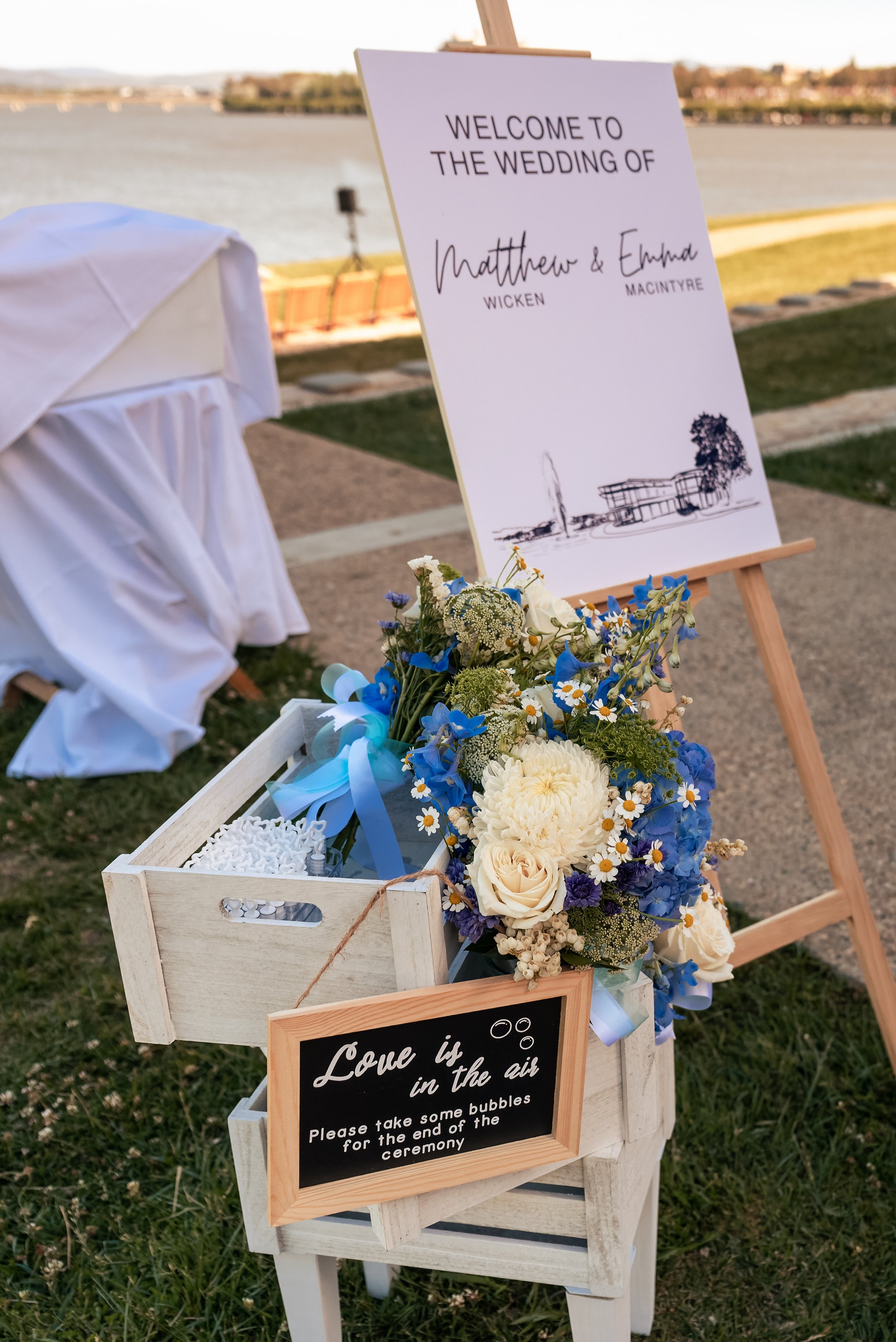 Welcome sign at a wedding by a river, with floral decoration and a basket of bubbles for guests to use during the ceremony.