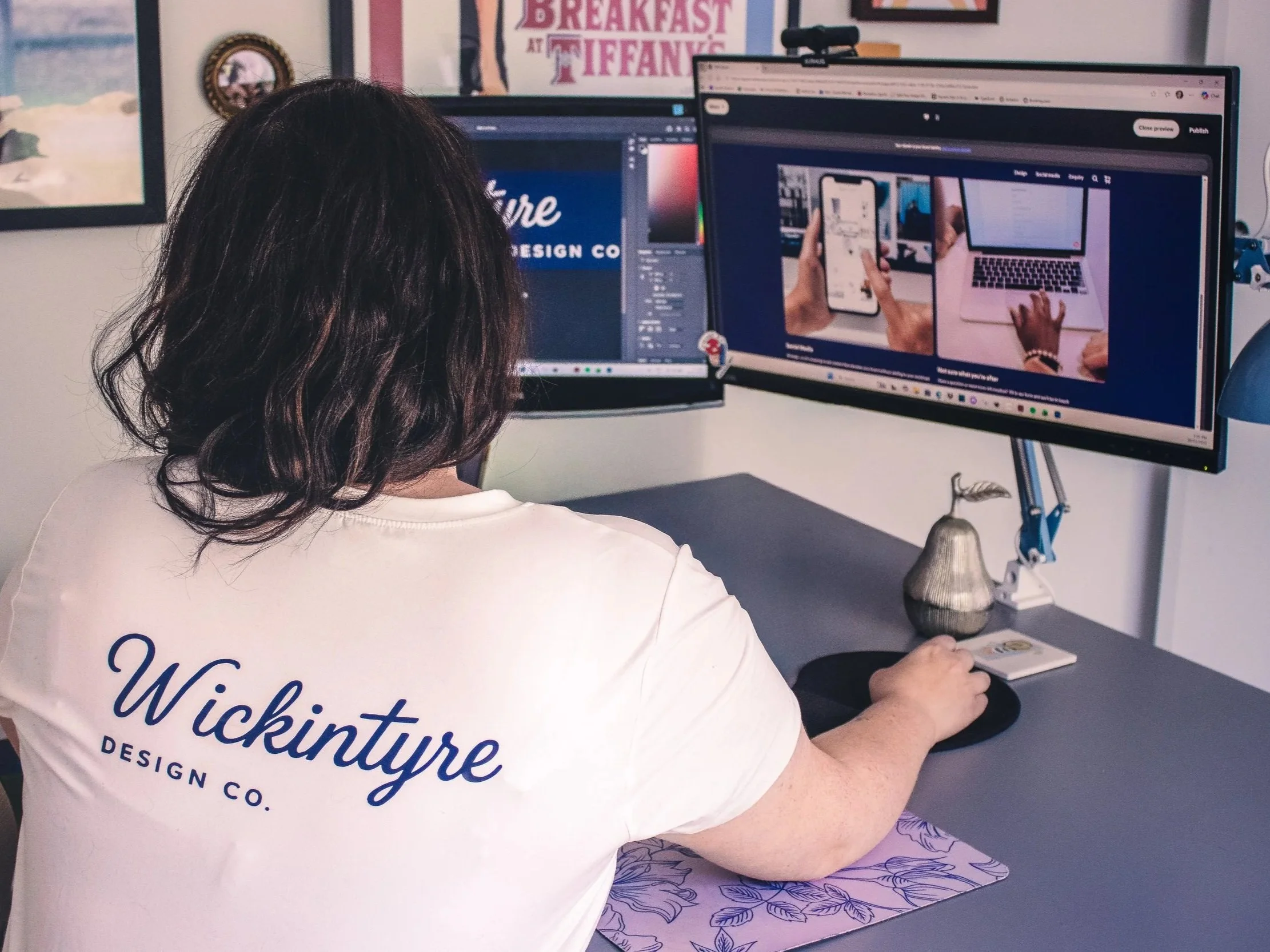 A woman with long dark hair seen from behind working on two computer monitors in an office. She is wearing a white shirt with blue text and is typing on a keyboard. The office wall has various posters, including one for breakfast at Tiffany's, and a small decorative object on the desk.