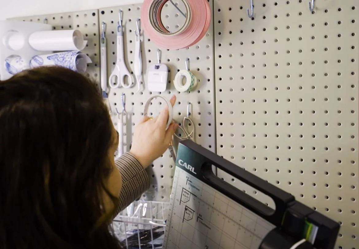 Person organizing a pegboard with various office supplies including tape, scissors, and rolls of paper or tape.