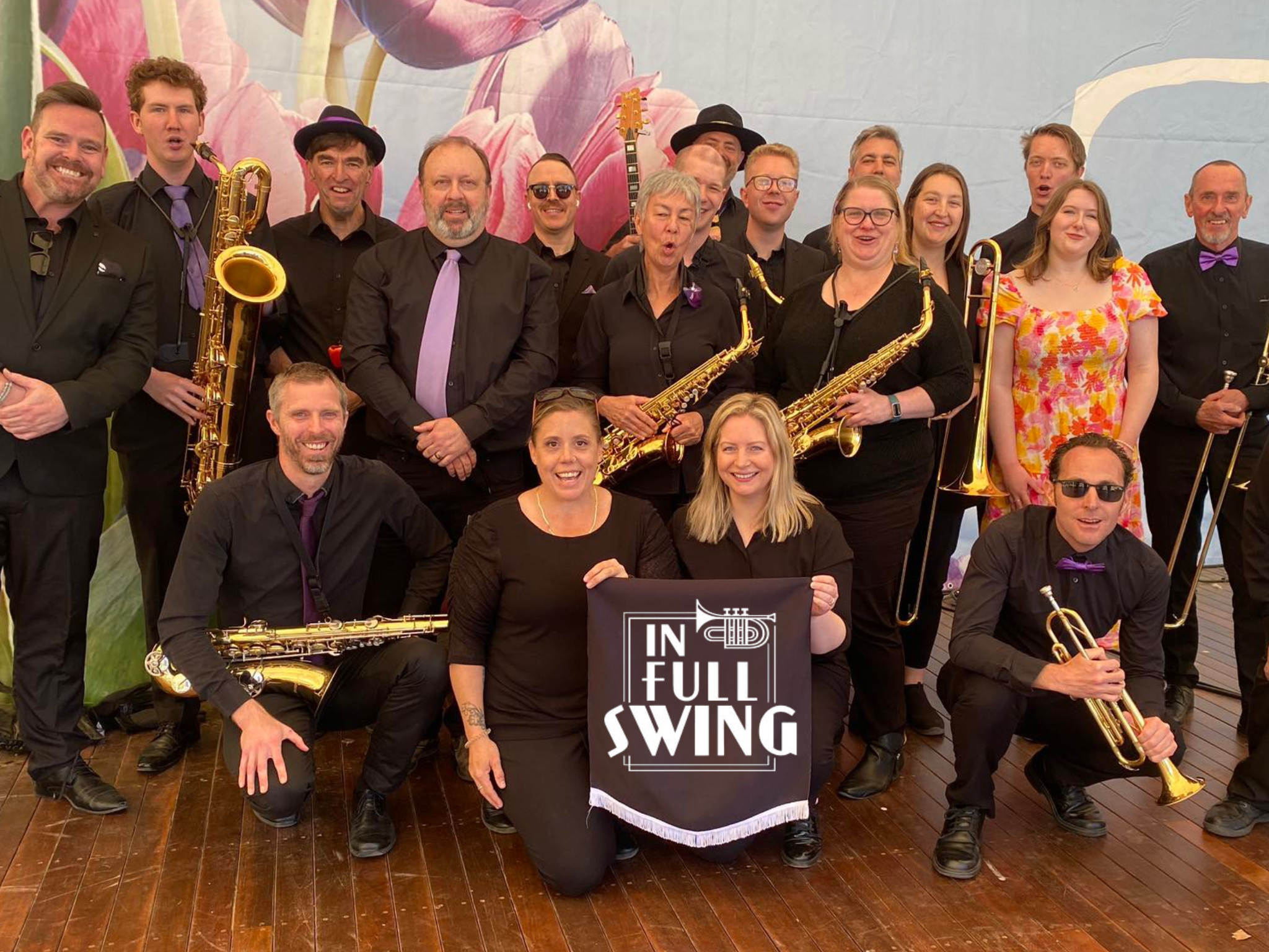 Group of ten musicians, mostly men, posing with musical instruments including saxophones, guitar, and clarinet, in front of a colorful backdrop with flowers. Some of them are smiling, and one woman is kneeling in front holding a flag or banner that reads 'FULL SWING'.