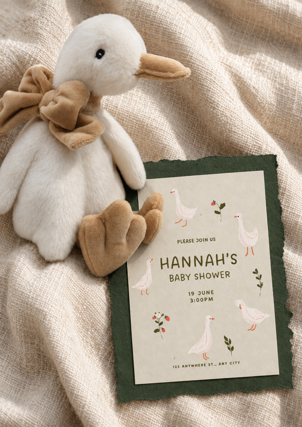A plush duck toy with a beige scarf around its neck and a beige bow, resting on a light beige textured fabric next to a baby shower invitation for Hannah, scheduled for June 19 at 3:00 PM, with illustrations of ducks and greenery on the invitation.