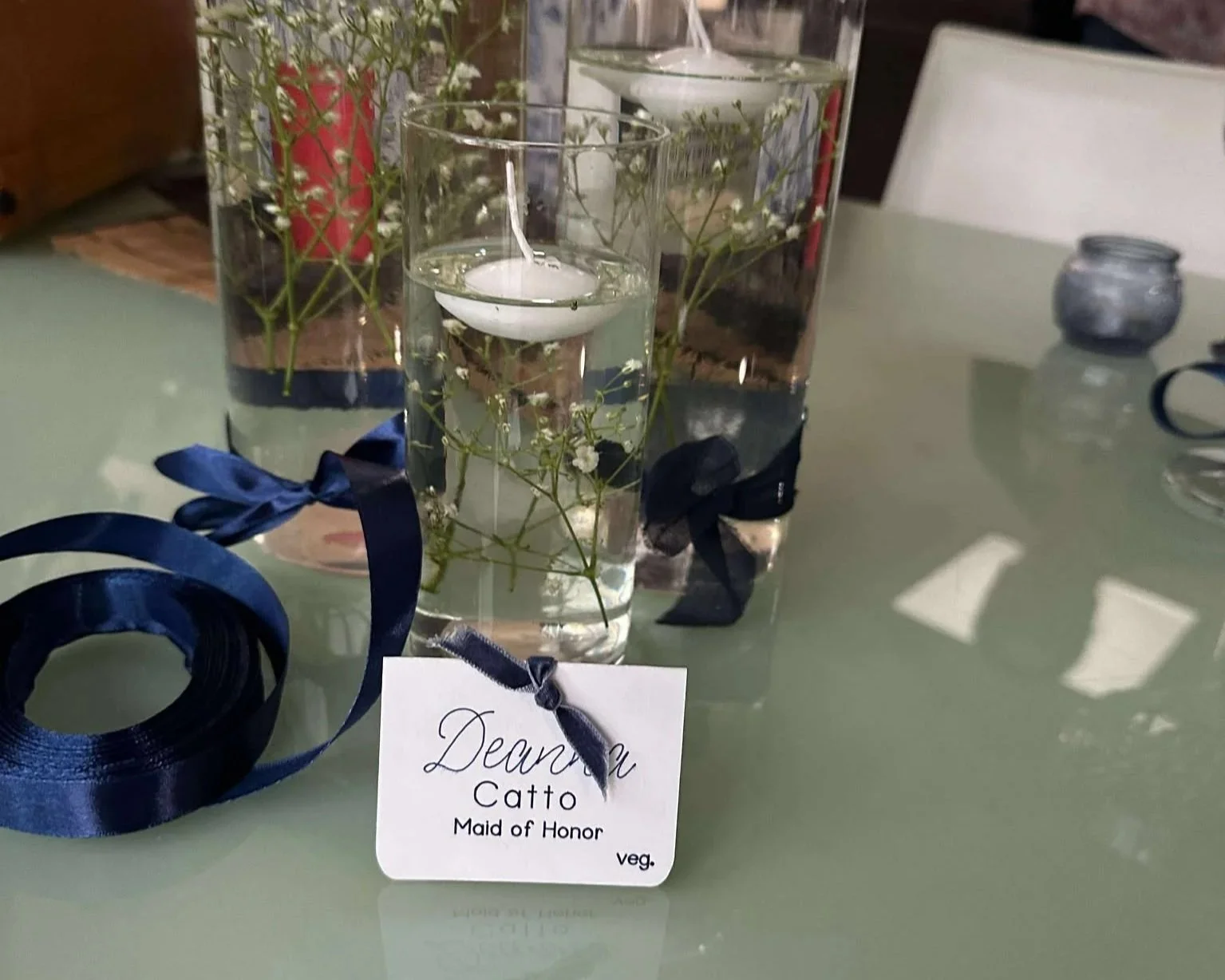 Decorative table centerpiece with three tall glass vases filled with water, floating candles, and greenery, accented with navy blue ribbons. A front tag reads, 'Dear You Catt0 Maid of Honor veg.'