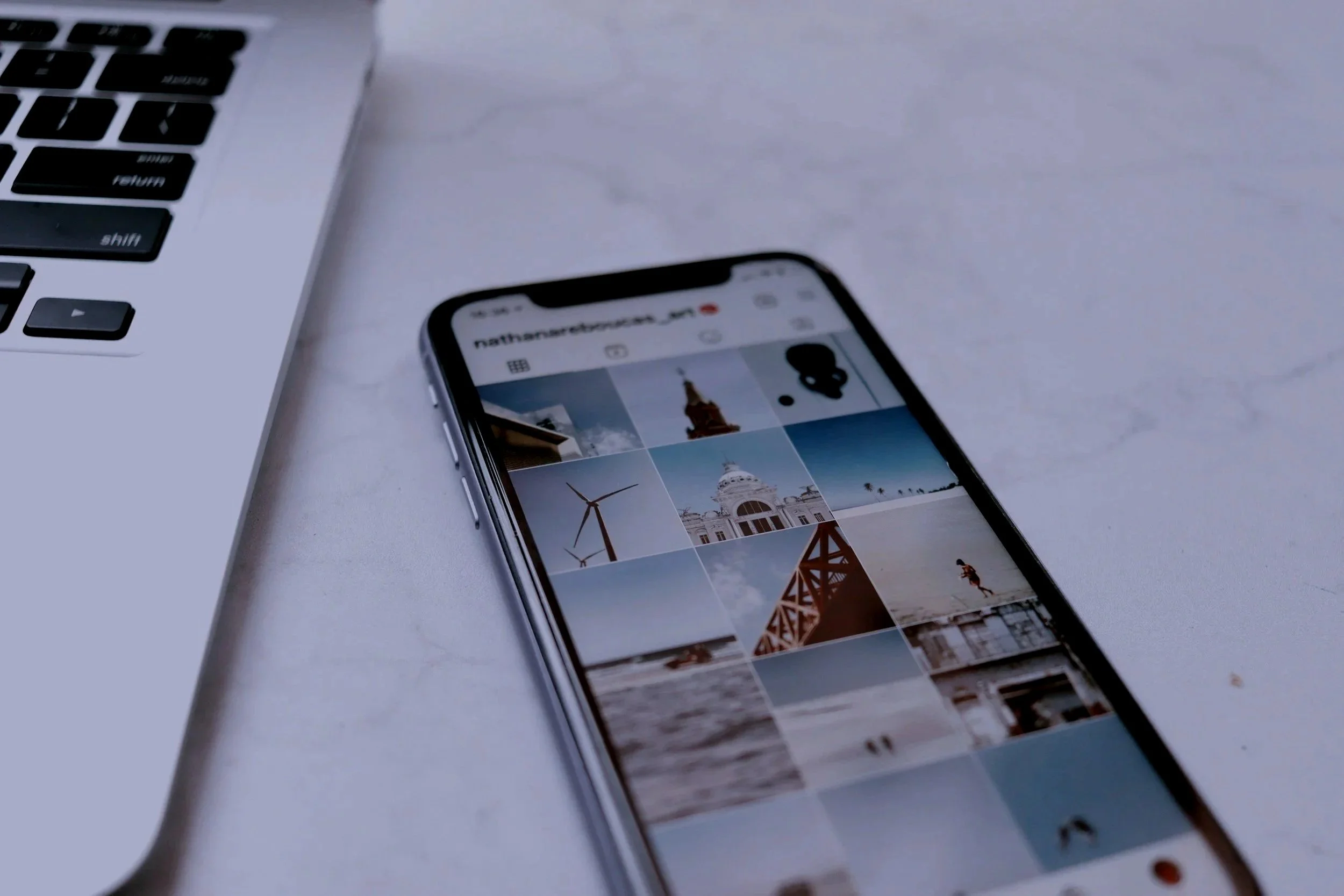 Close-up of a smartphone displaying a grid of travel photos, next to a silver laptop on a white desk.