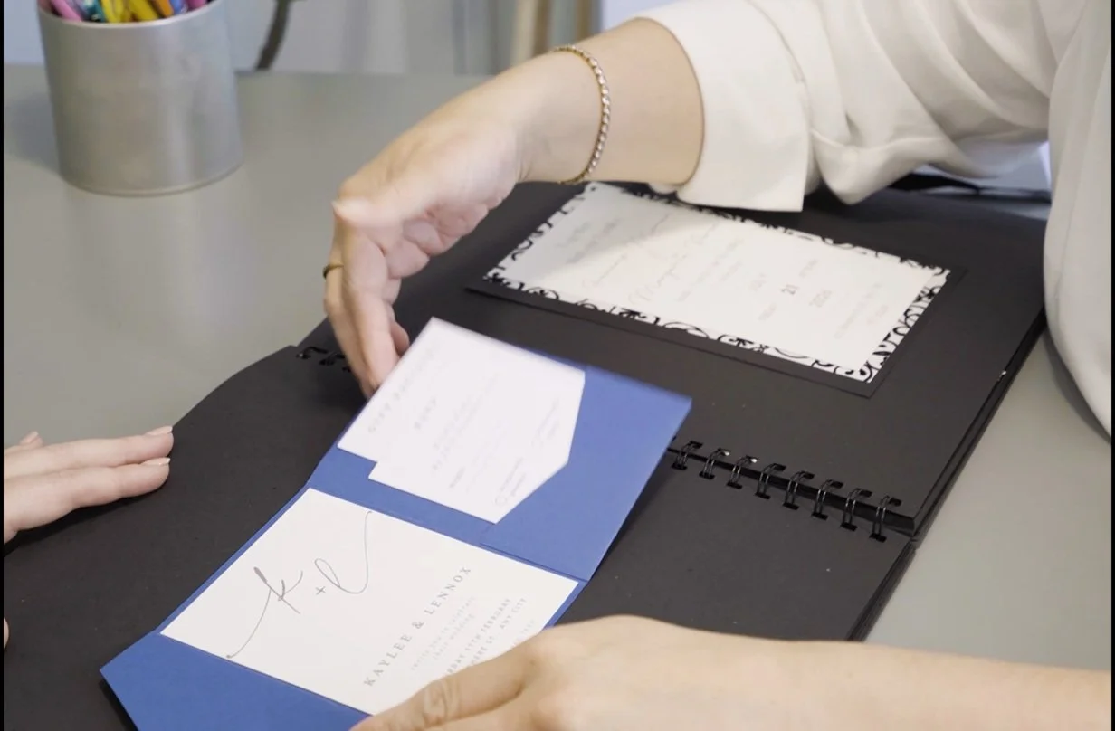 Person showing wedding invitation and RSVP card to another person on a black presentation board.