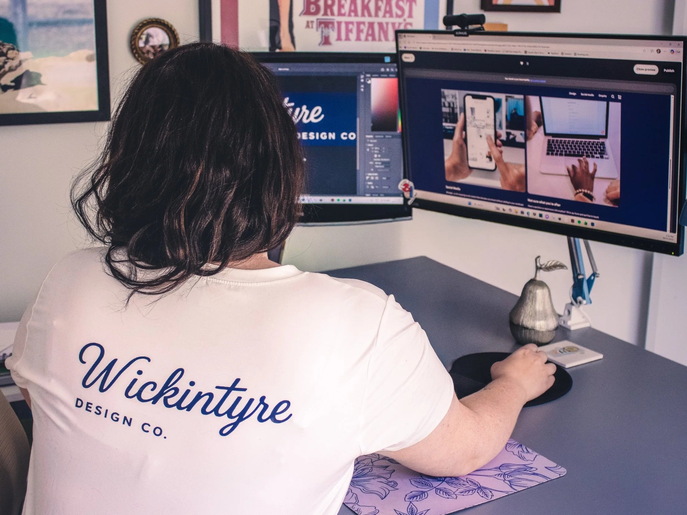 A woman with dark hair sitting at a desk, looking at dual computer monitors. She is wearing a white t-shirt with blue lettering that says 'Wickington' and some other text. The monitors display various images, including a website and a photo of a person holding a phone and typing on a laptop. There is a framed picture and decorative items on the wall and desk.