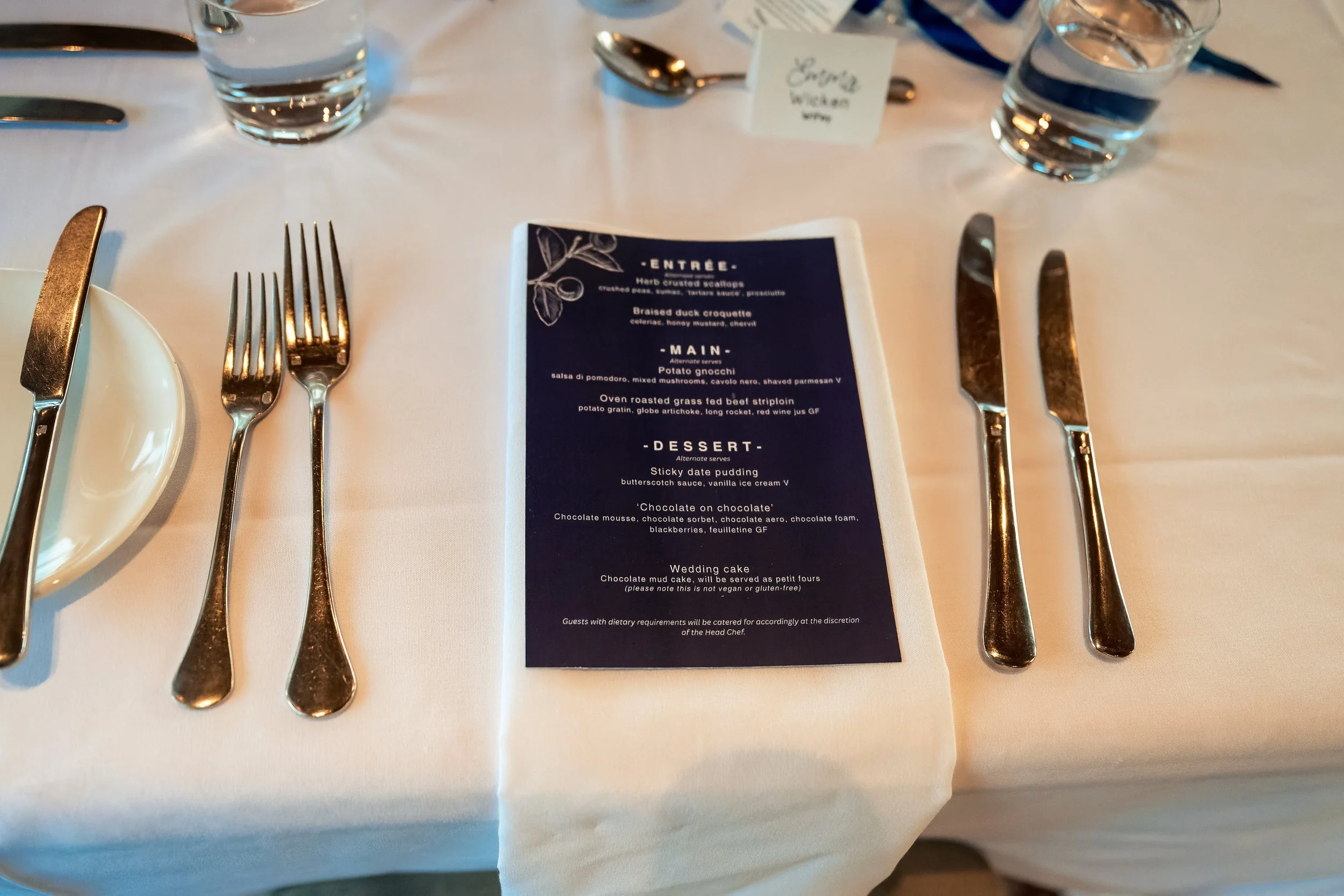 A white table setting with a printed menu card, three water glasses, and silverware including a knife, a fork, and a spoon. The menu features various food options and dessert items.