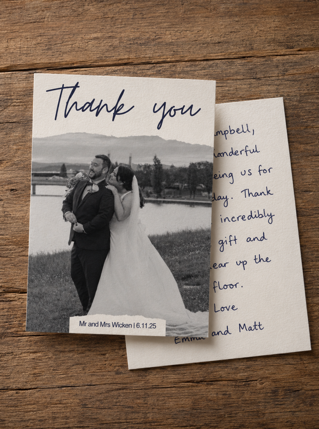 A black-and-white wedding thank you card featuring a photo of a bride and groom standing outdoors near a body of water, with mountains in the background. The card is on a wooden surface, with the front visible and the back partially showing handwritten message.