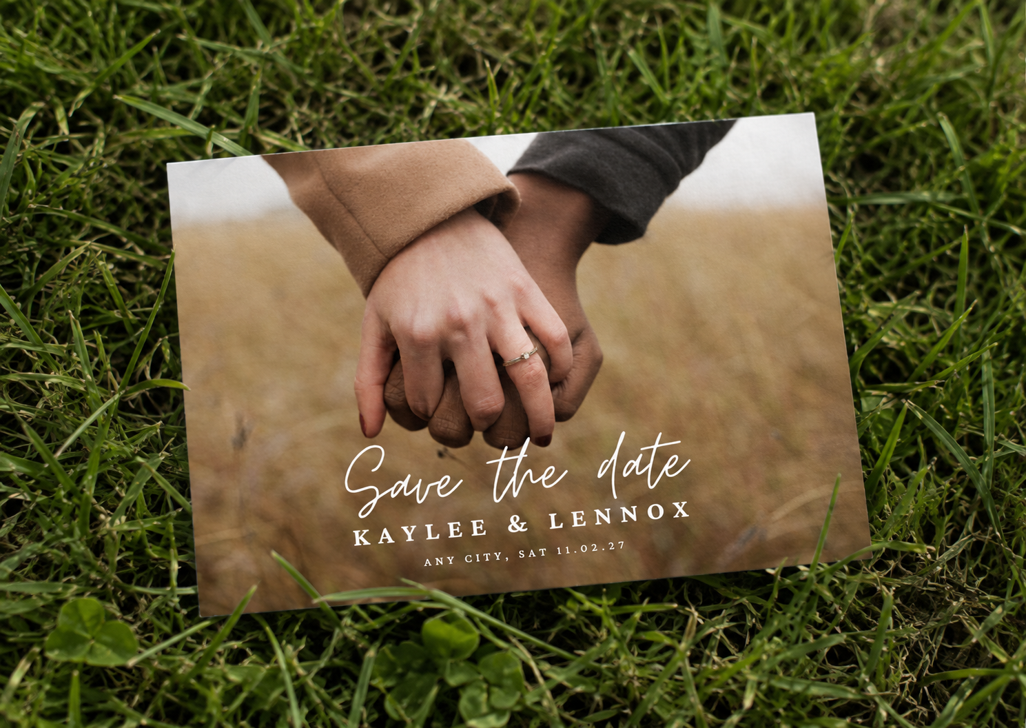 Save the date card featuring a couple holding hands, with wedding details beneath, laid on grass.