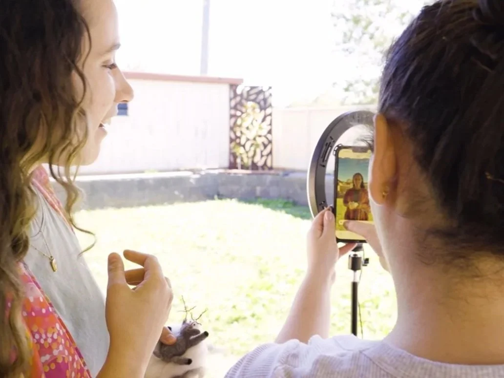 Two women taking a selfie of each other outdoors, one holding the phone and the other smiling. The scene is set in a sunny backyard with a fence and grass.