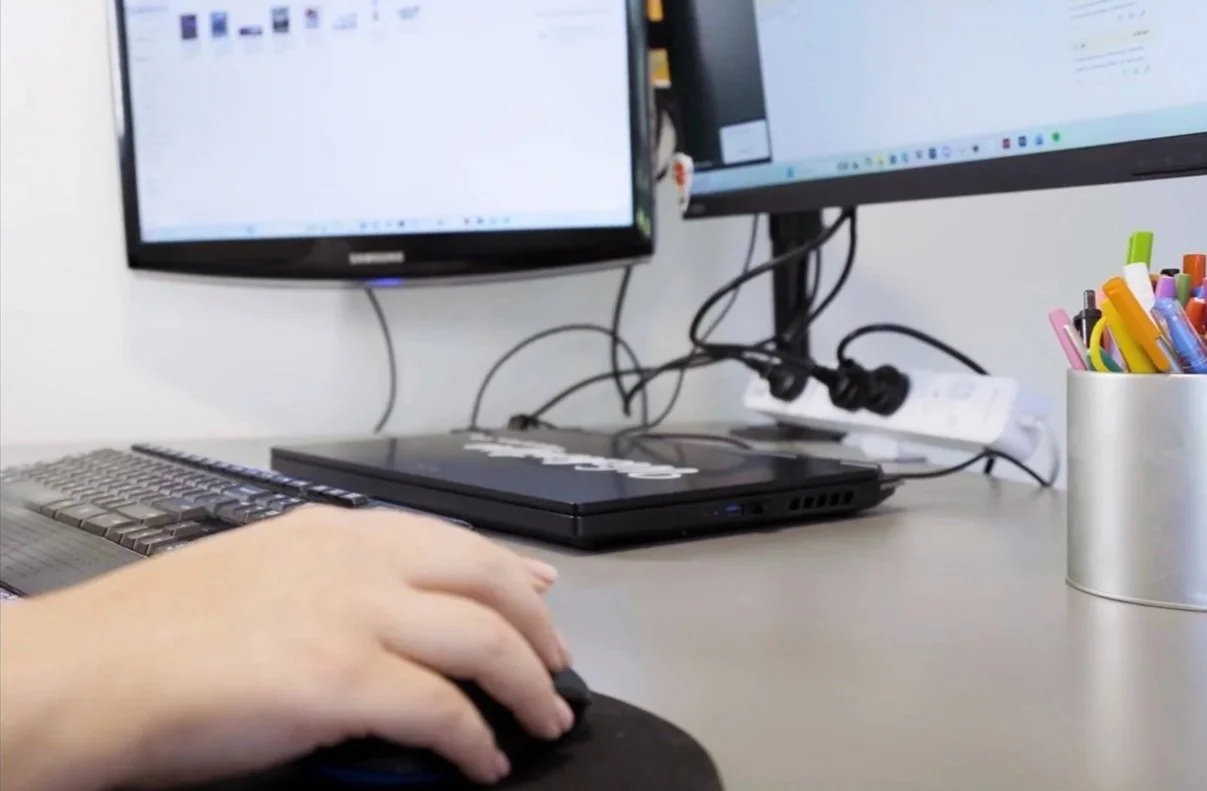Close-up of a person's hand on a computer mouse, with dual computer monitors, a keyboard, a closed laptop, a white power strip, and a container filled with colorful pens on a desk.