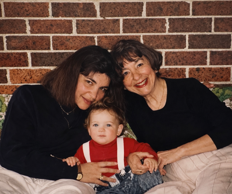 Three generations of women, a young woman, an elderly woman, and a toddler, sitting on a couch in front of a brick wall, smiling and holding hands.