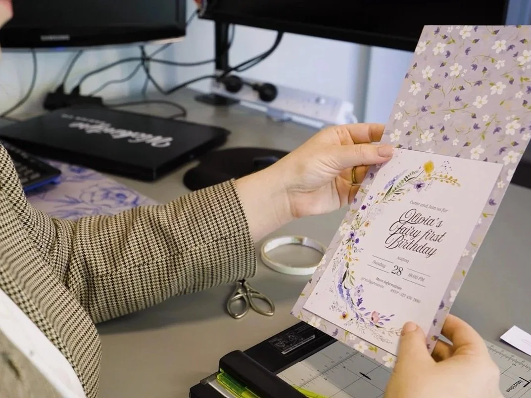 Person holding a floral birthday invitation card with a wedding-themed design, on a desk with scissors, a cutter, and other office supplies.