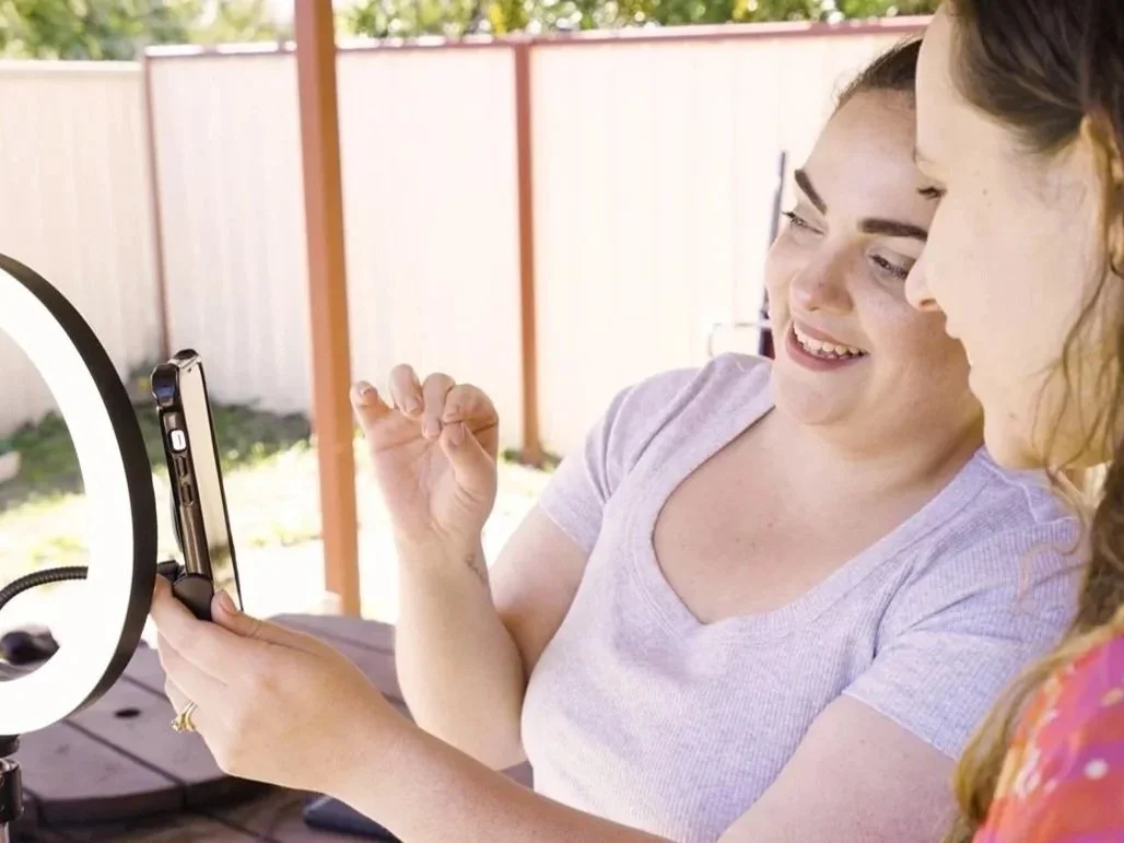 Two women sitting together outside, smiling and looking at a smartphone held by one of them, with a ring light nearby.
