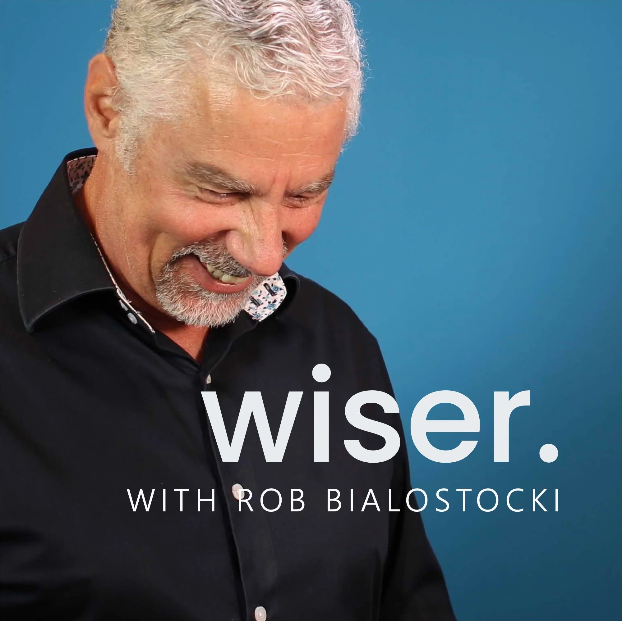 Close-up of a smiling man with white hair and a beard, wearing a black shirt with a colorful collar, against a blue background with partially visible white text that reads 'Wisdom' and 'With Rob Bialostocki'.