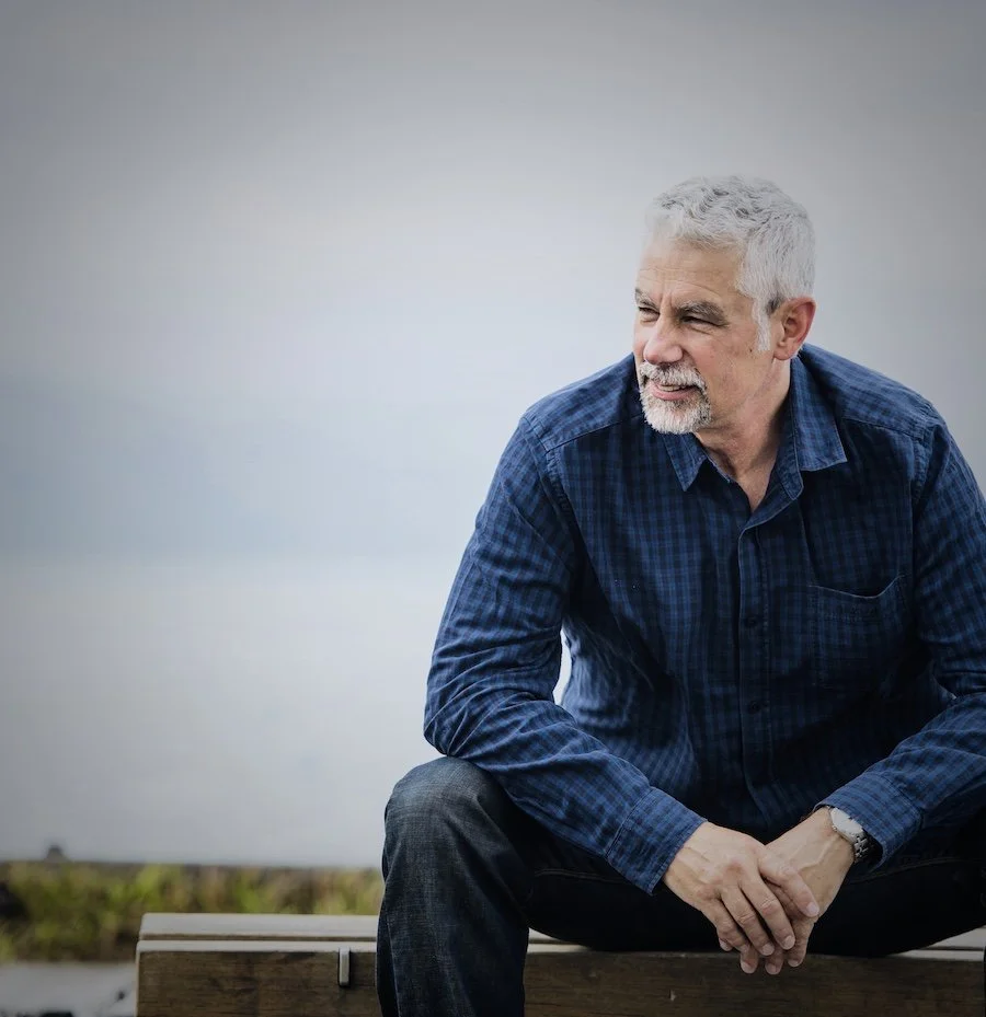 An older man with white hair and a beard sitting on a bench outdoors, wearing a blue plaid shirt and dark jeans, looking thoughtful.