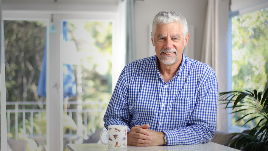 A smiling older man with gray hair and a beard sitting at a table in a bright kitchen or dining area, wearing a blue and white checkered shirt, with a coffee mug on the table and a window with sunlight and greenery outside in the background.