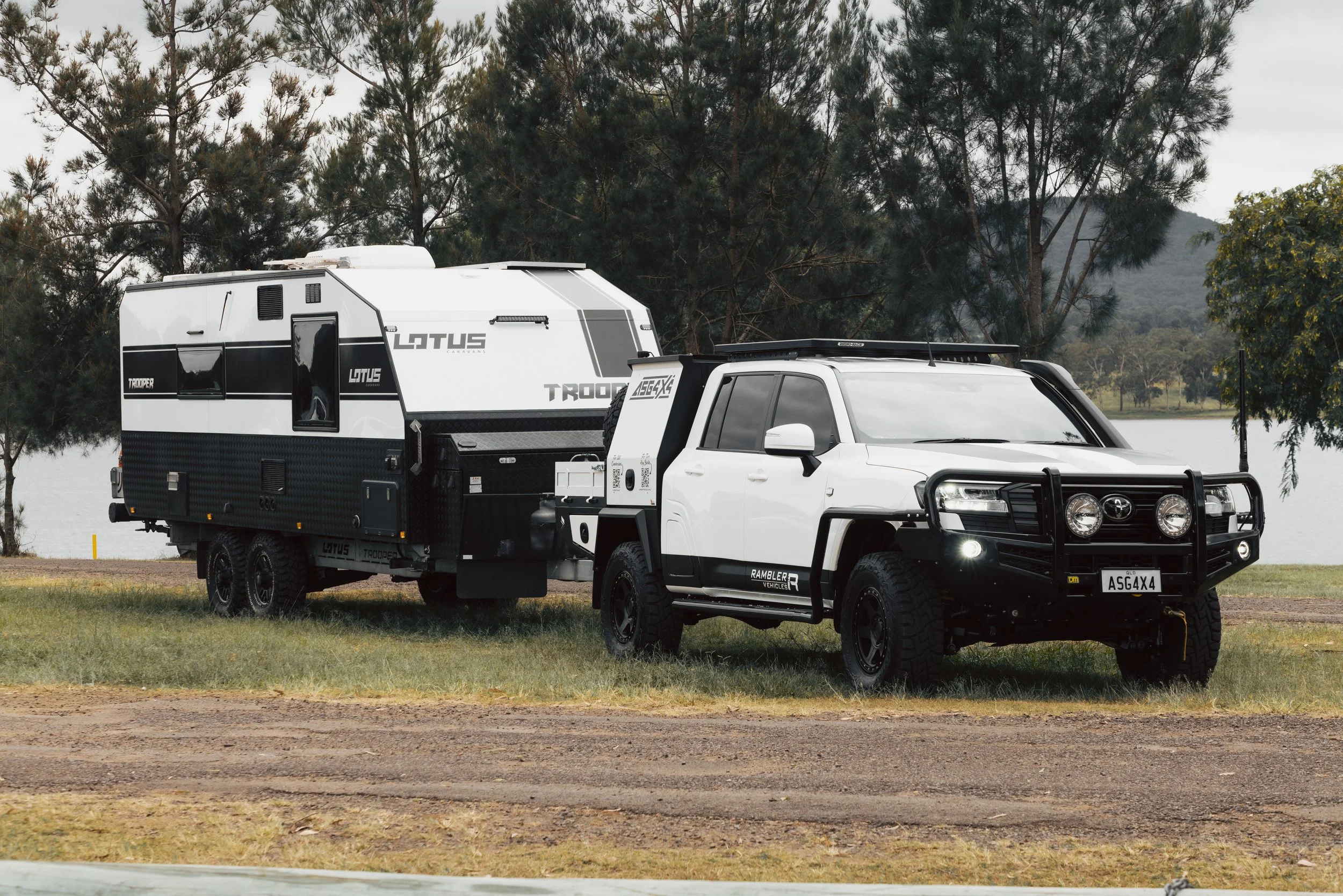 A white pickup truck with off-road tires, a black front bumper guard, and a rooftop antenna parked on grass near a lake. The truck is hauling a large, white camper trailer with black accents surrounded by tall trees with a mountainous backdrop.