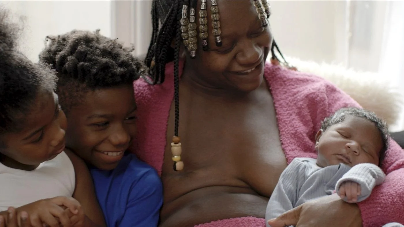 A woman with braided hair and beads, wrapped in a pink blanket, smiling at a newborn baby she is holding. Two children are beside her, smiling and looking at the baby.
