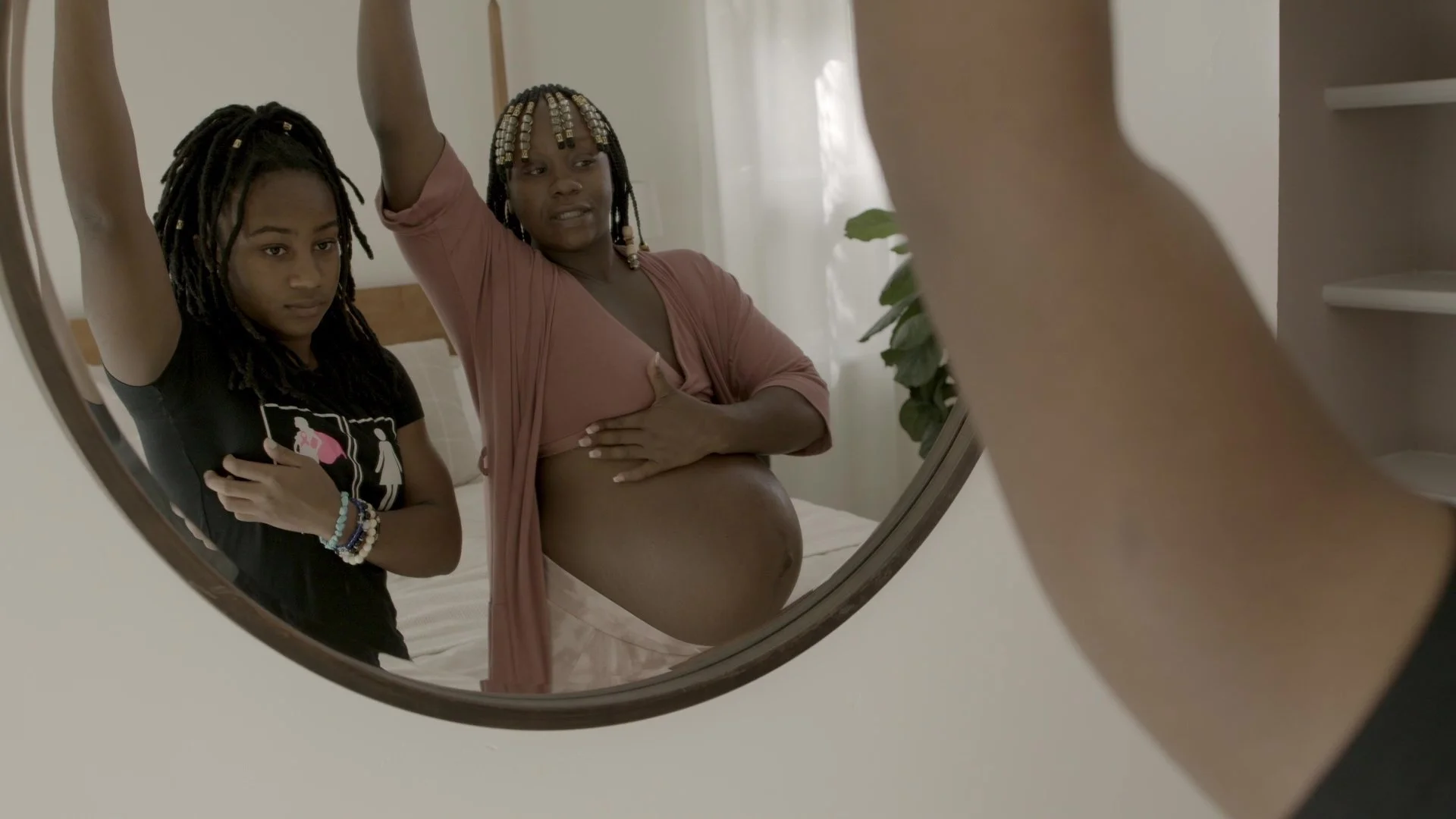 Reflection of two women, one pregnant, taking a mirror selfie in a bedroom with white walls, bed, and a potted plant.