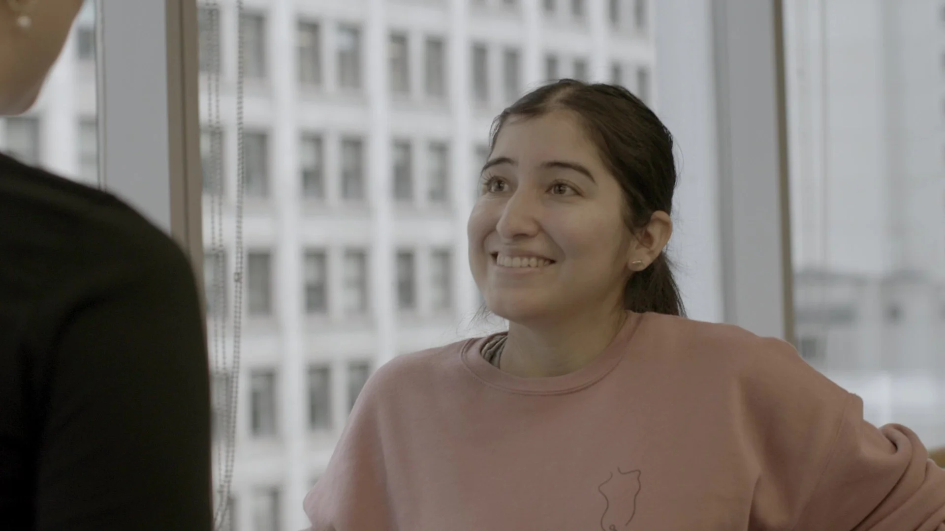 A young woman with dark hair smiling and looking at a person in front of her, indoors with a view of tall buildings through large windows.