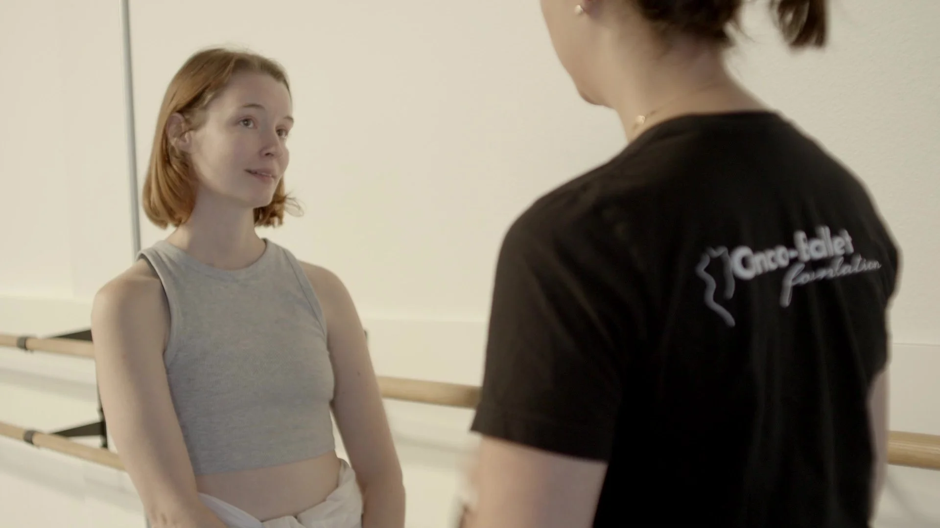 A young woman with red hair wearing a gray tank top and white pants standing in front of a ballet barre, engaging in conversation with an instructor wearing a black shirt with the Onco-Teacher Foundation logo.