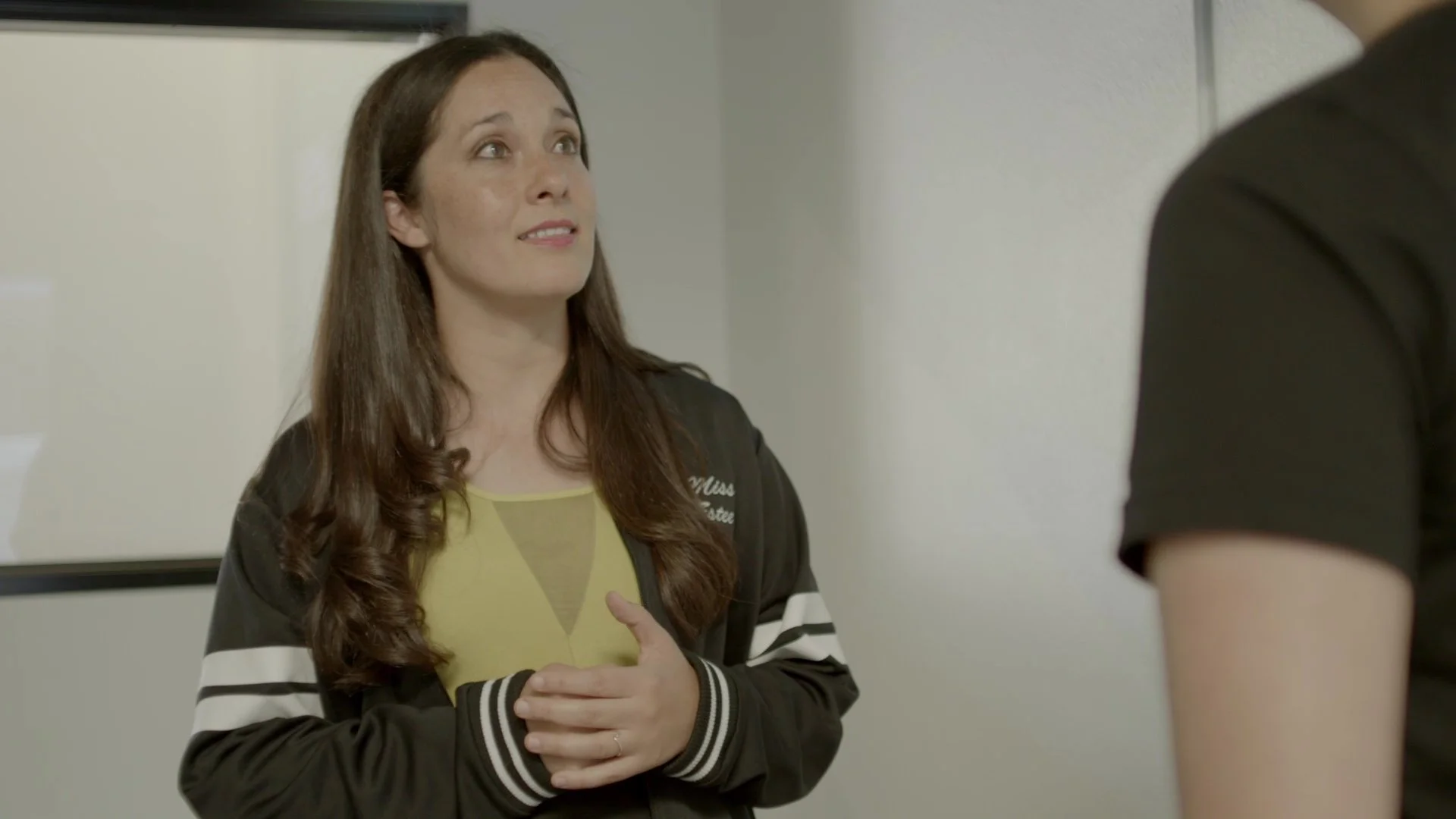 A woman with long brown hair wearing a black and white jacket and light green top, talking to a person in a black t-shirt, in an indoor setting.