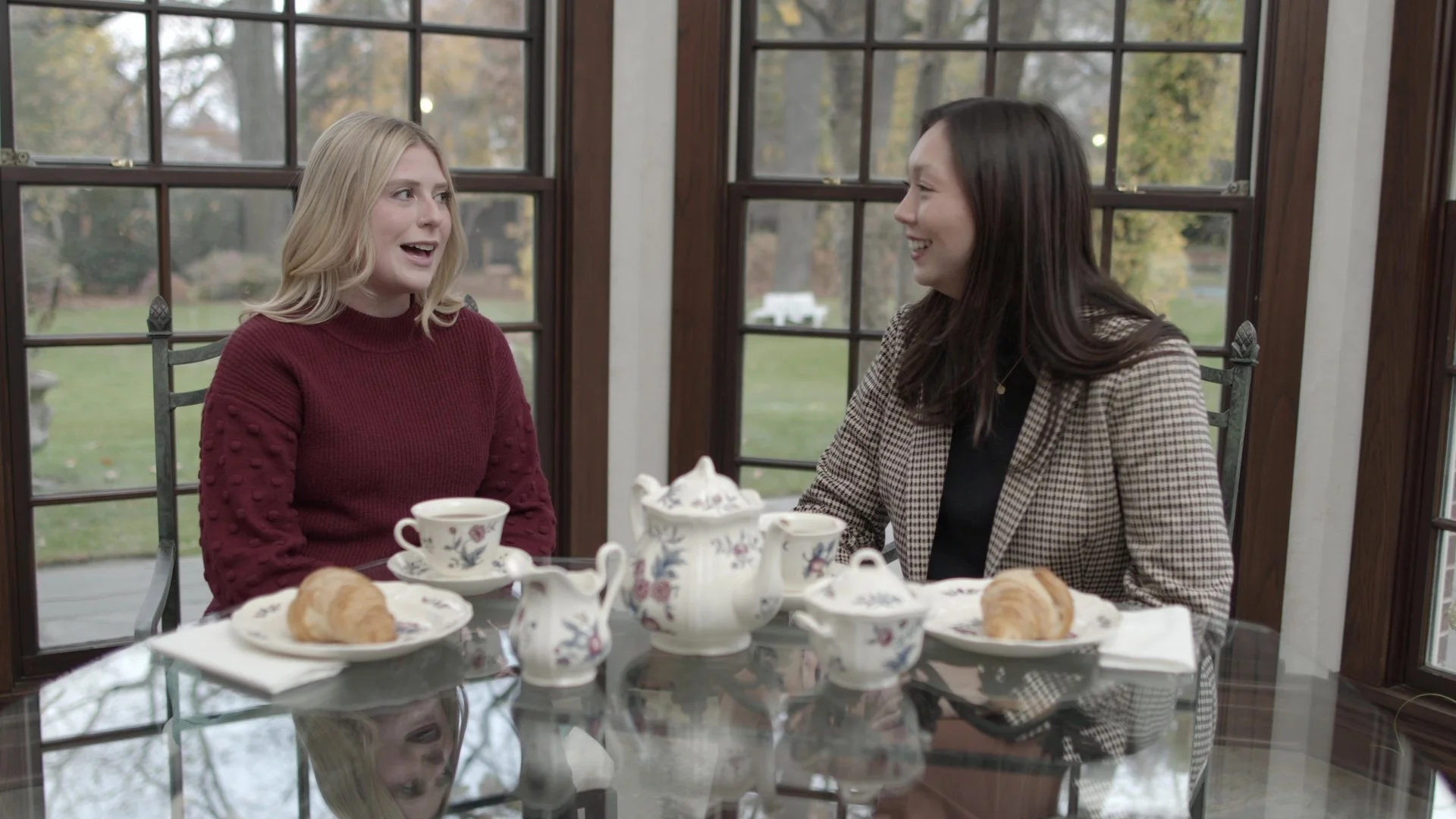 Two women sitting at a dining table with tea cups, teapot, and pastries, having a conversation in a sunlit room with large windows showing an outdoor garden.