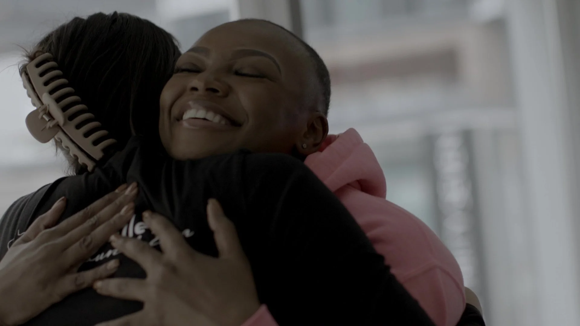 Two women hugging each other with smiles, one woman wearing a pink hoodie and the other having a hair roller, in a bright indoor setting.