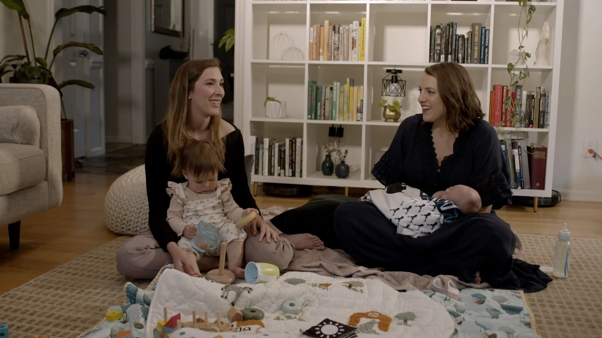 Two women and two babies sitting on a rug in a living room, engaging with each other and toys, with a bookshelf behind them.