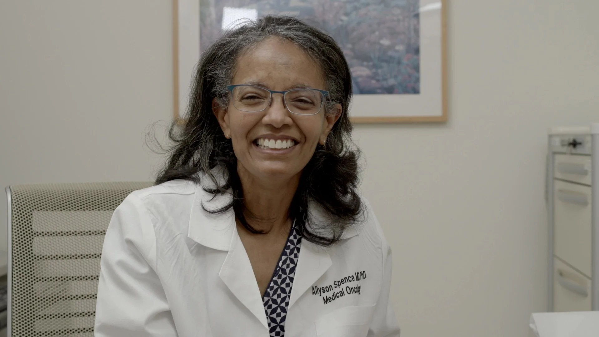 A smiling African-American woman with gray hair, wearing glasses and a white medical coat, sitting in an office. Behind her, there is a framed picture on the wall and a set of drawers to the right.