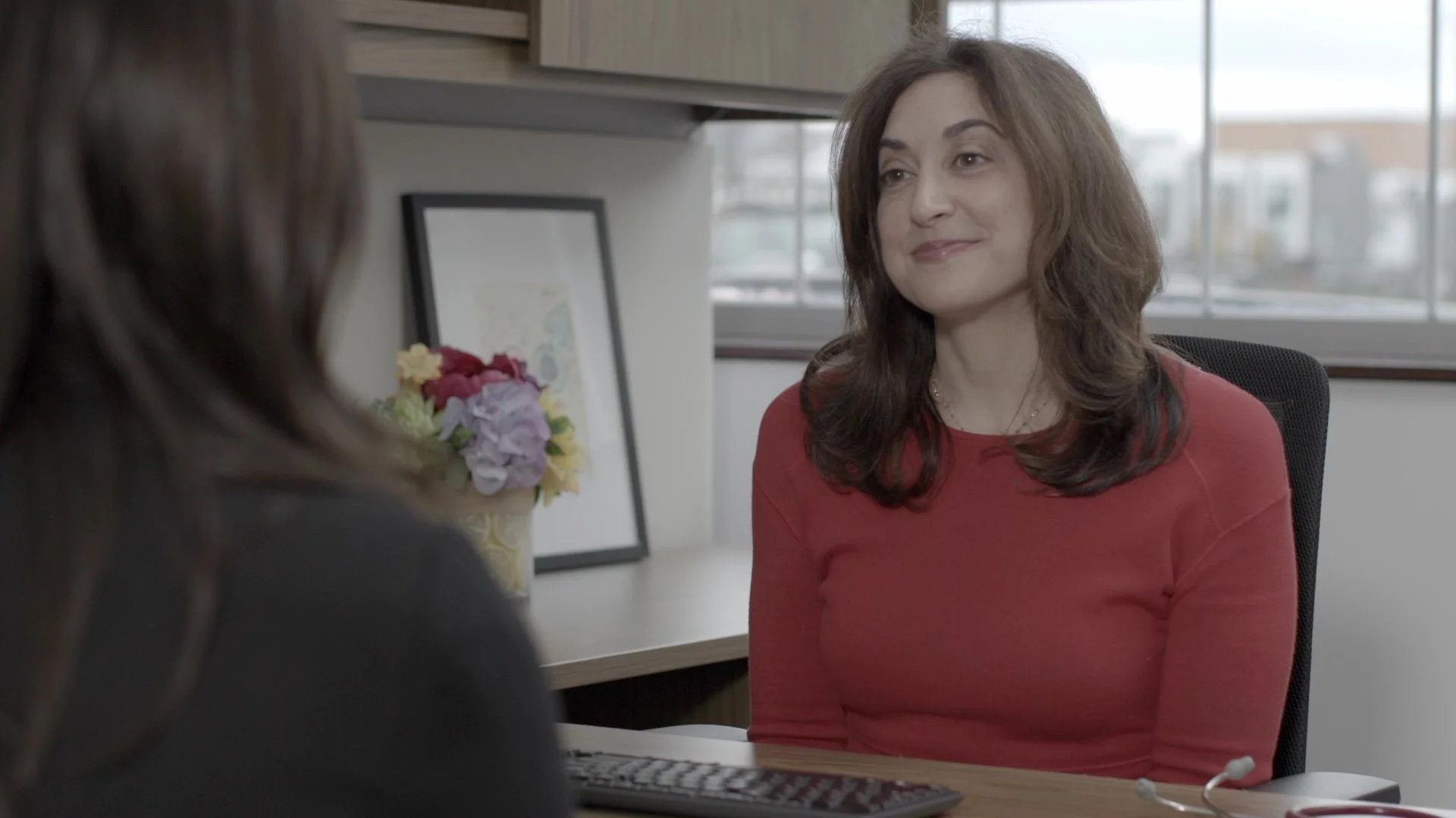 A woman with shoulder-length brown hair wearing a red top sitting at an office desk, having a conversation with another person whose back is facing the camera. Behind her, there is a window with a view of buildings, a framed picture, and a vase of flowers on the desk.