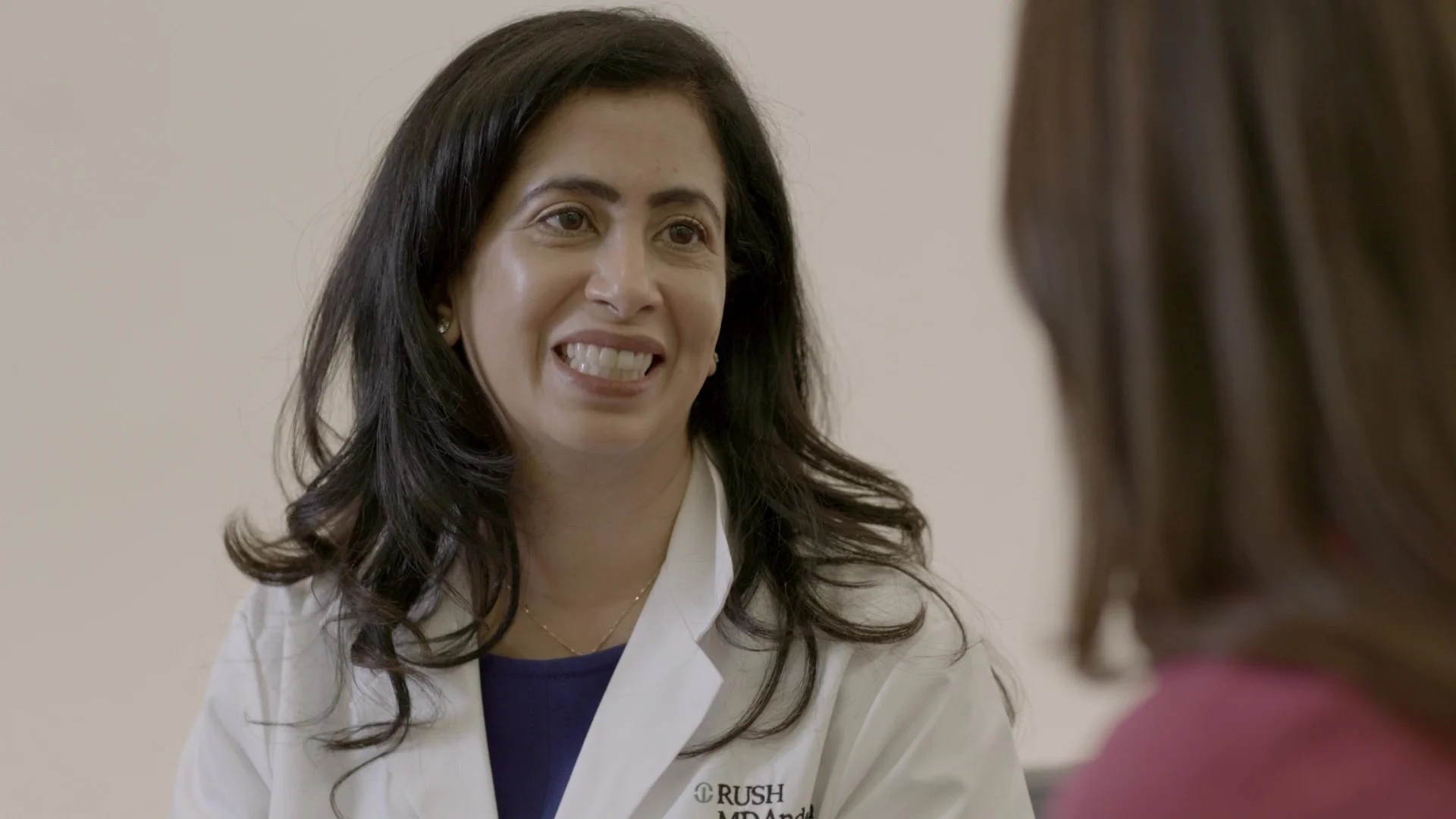 A woman with dark hair and a white coat smiling while talking to a person whose back is visible in the foreground.