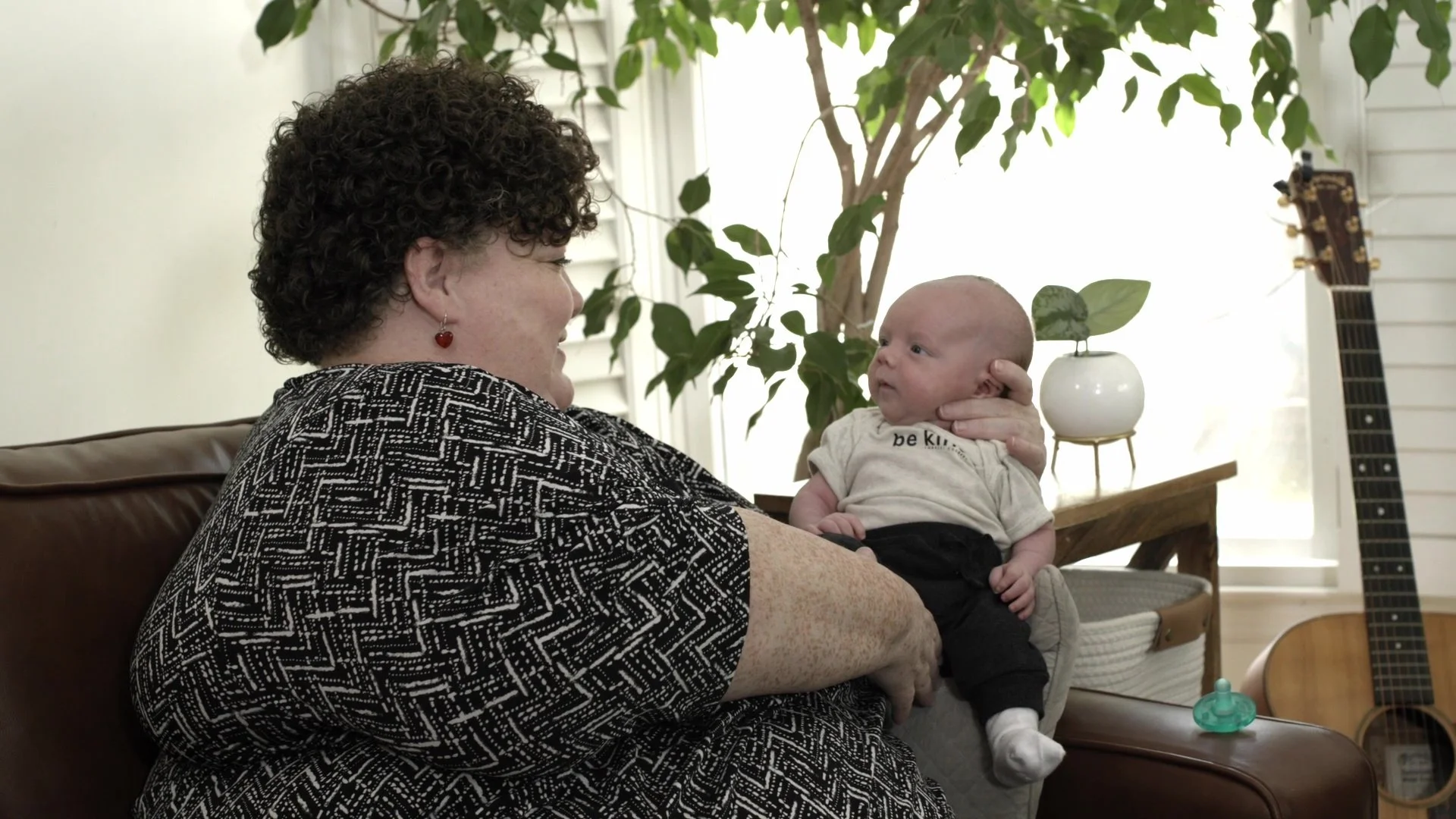 An older woman with curly hair and red earrings holds a baby boy on her lap indoors. The woman has a patterned black and white top and is smiling at the baby. The baby looks back at her with curiosity and has a shaved head, wearing a light-colored shirt and dark pants. In the background, there is a large leafy plant, a guitar, a small white planter on a table, and chair.