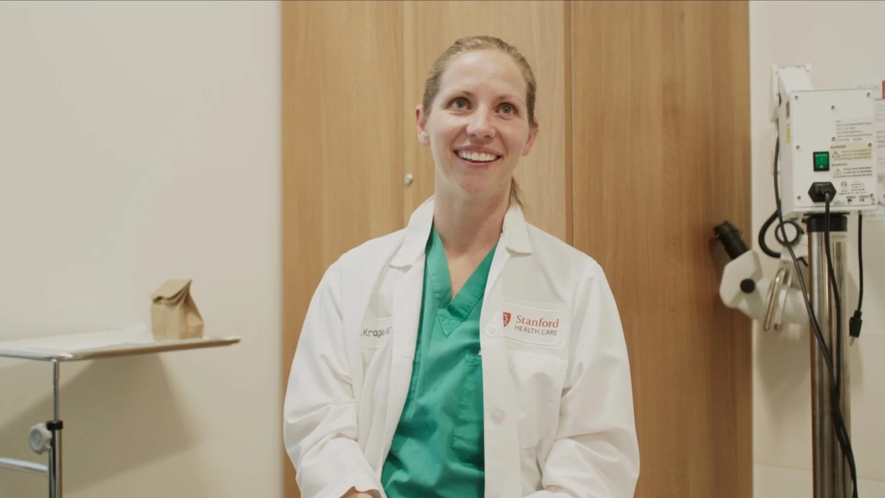 A smiling female healthcare professional in a white lab coat and green scrubs standing in a medical setting near medical equipment and a wooden wall.