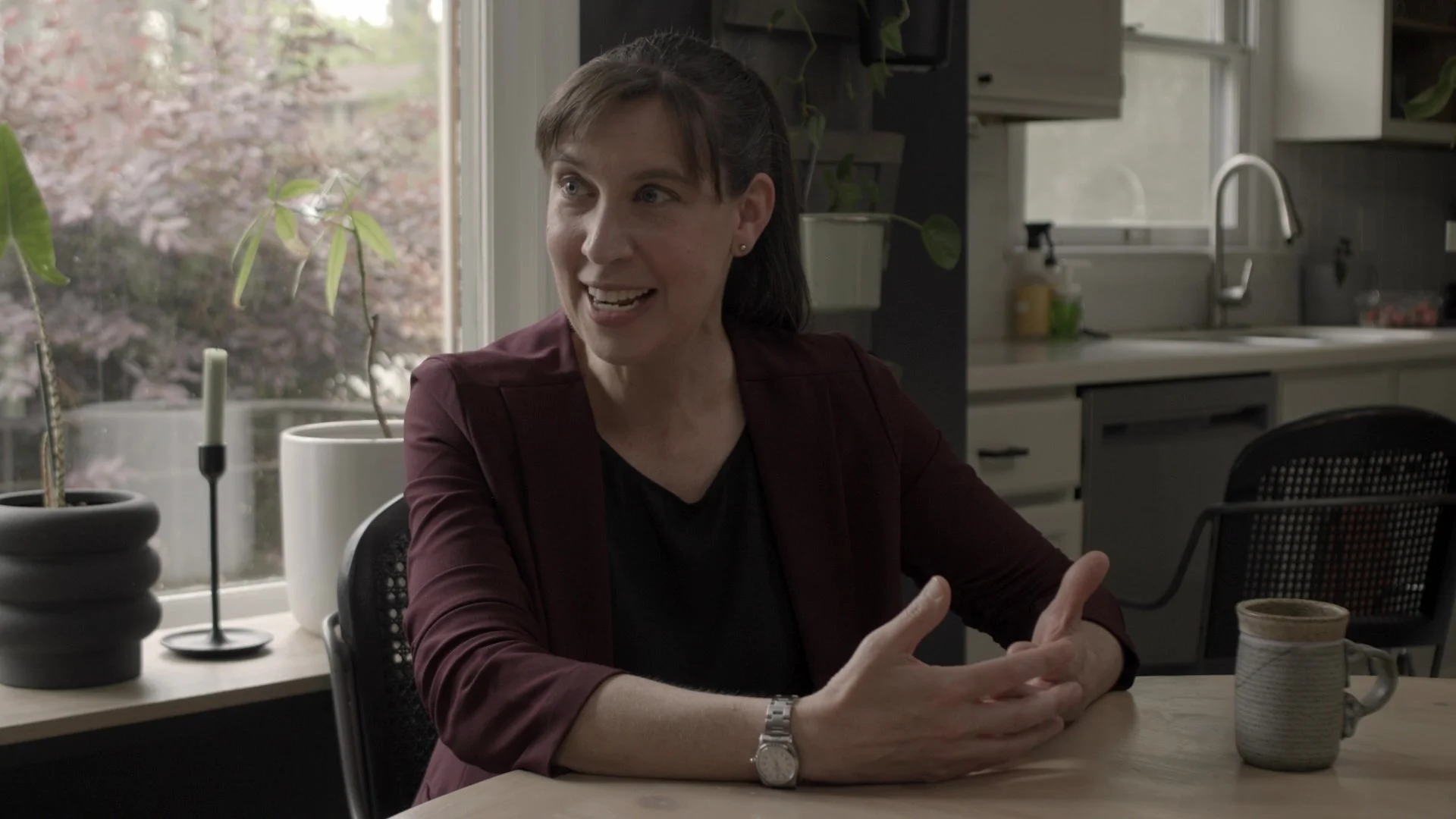 A woman with brown hair, wearing a dark blazer and a watch, is sitting at a table indoors, smiling and talking. There is a large window behind her with a view of trees outside, and on the table are a mug and some decorative items.