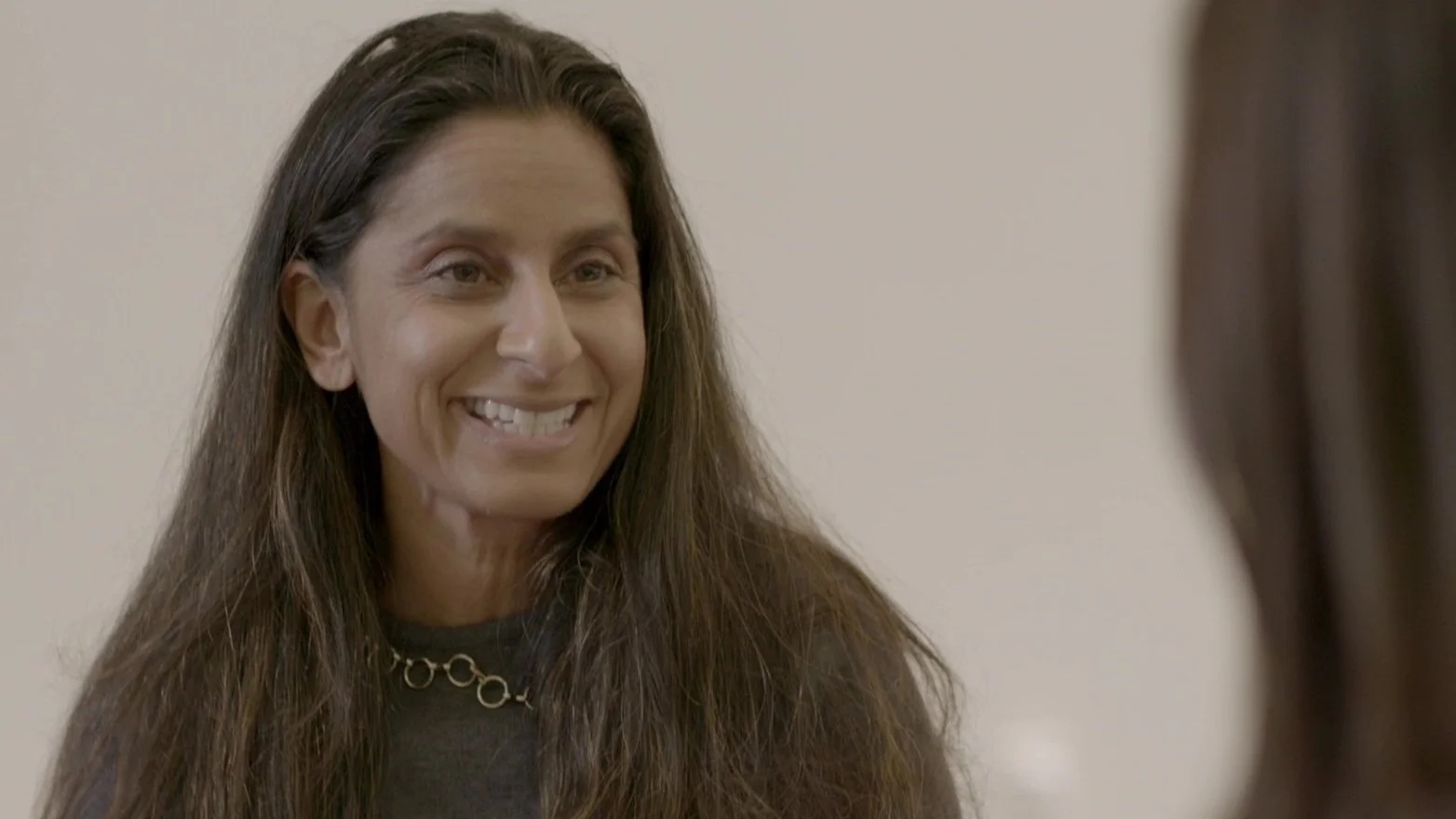 A woman with long brown hair smiling and talking to another person whose back is visible.