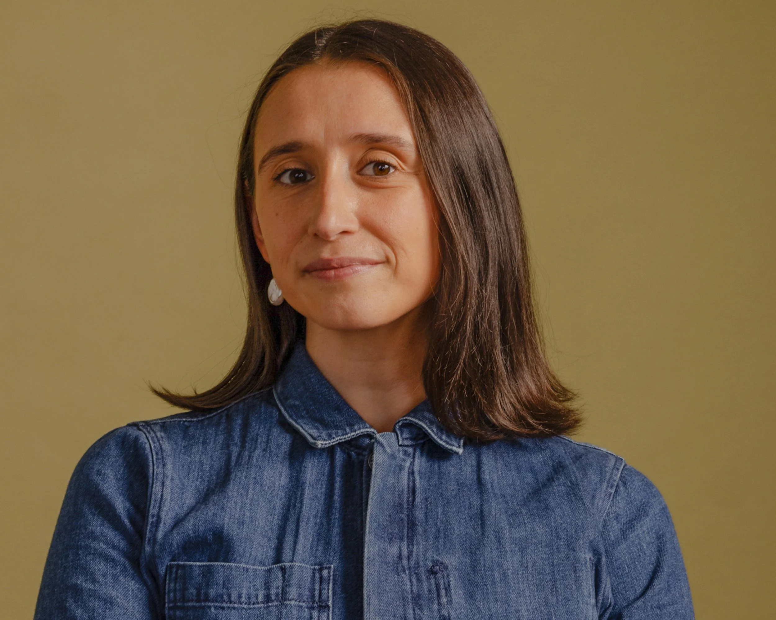 A young woman with brown hair and brown eyes, wearing a blue denim shirt, standing in front of a yellow background. She is smiling slightly and looking at the camera.