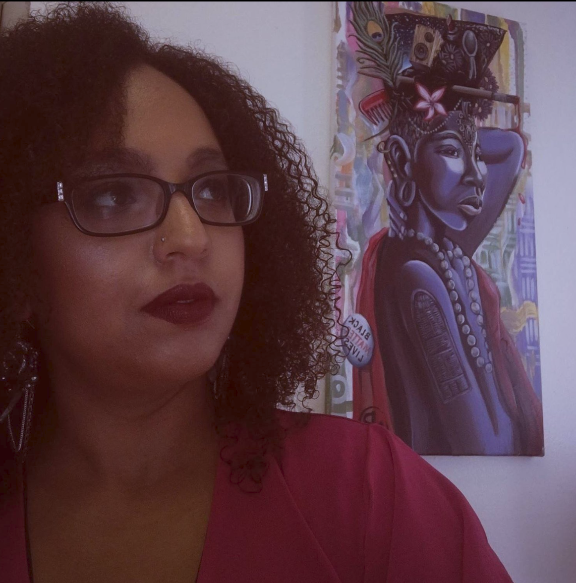 A beautiful African American woman with curly hair and glasses looks off into the distance. An African painting is in her background.