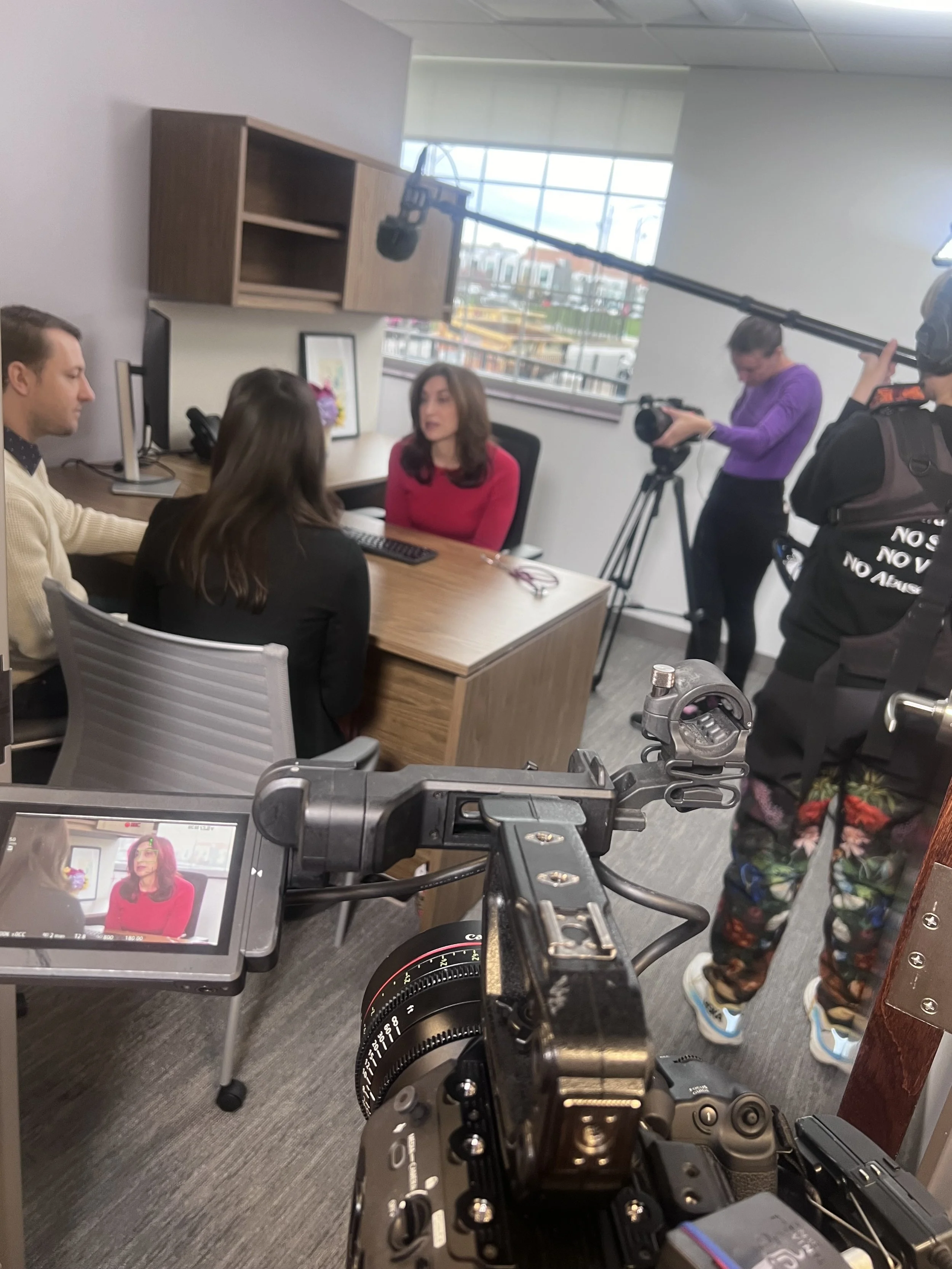 A behind-the-scenes look at a video shoot in an office, with a woman in a red top being filmed, a cameraman recording her, a crew member managing equipment, and a monitor displaying the woman's image.