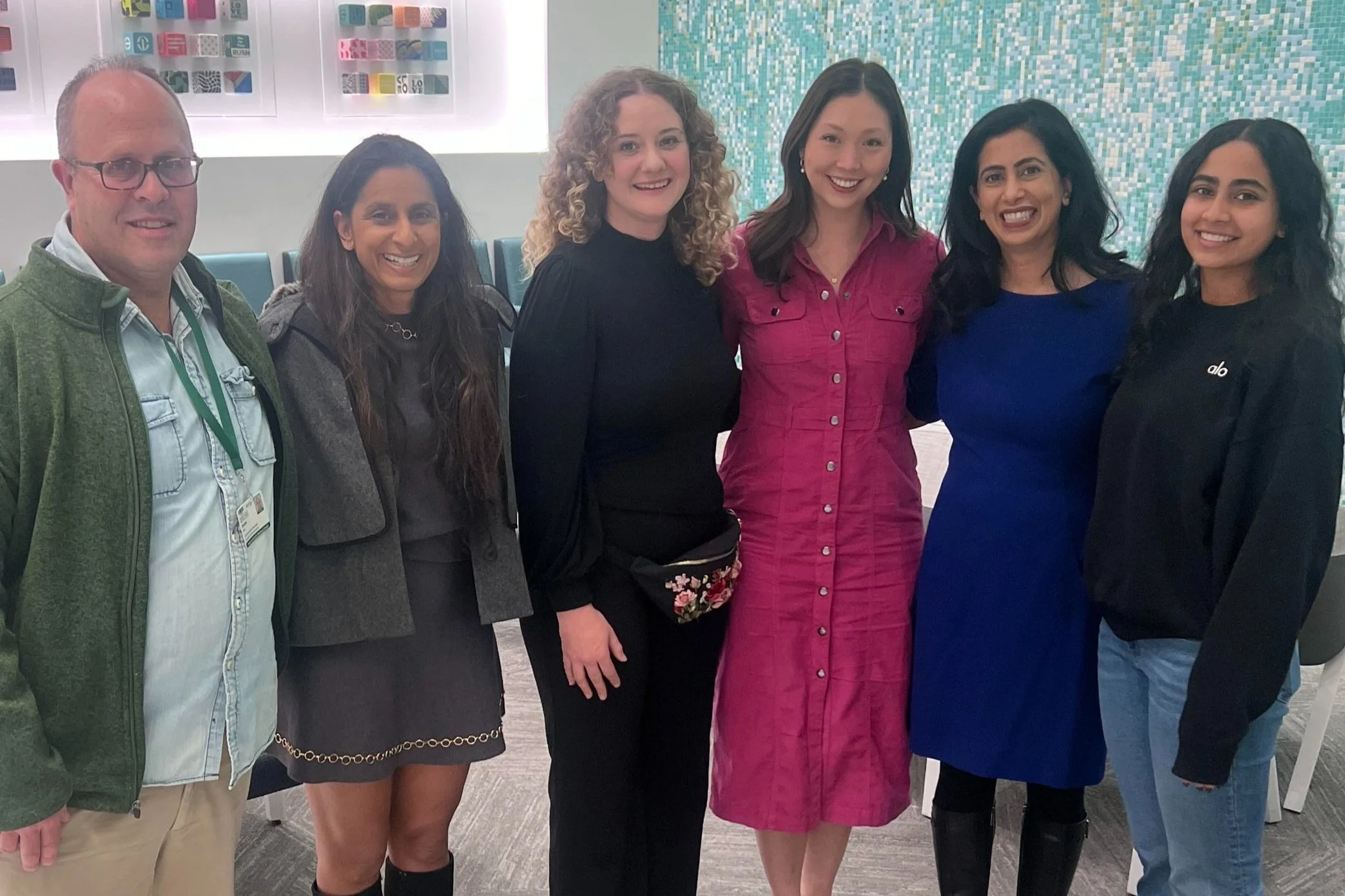 Group of six diverse women and one man standing together, smiling in an indoor setting with artwork and a mosaic wall in the background.