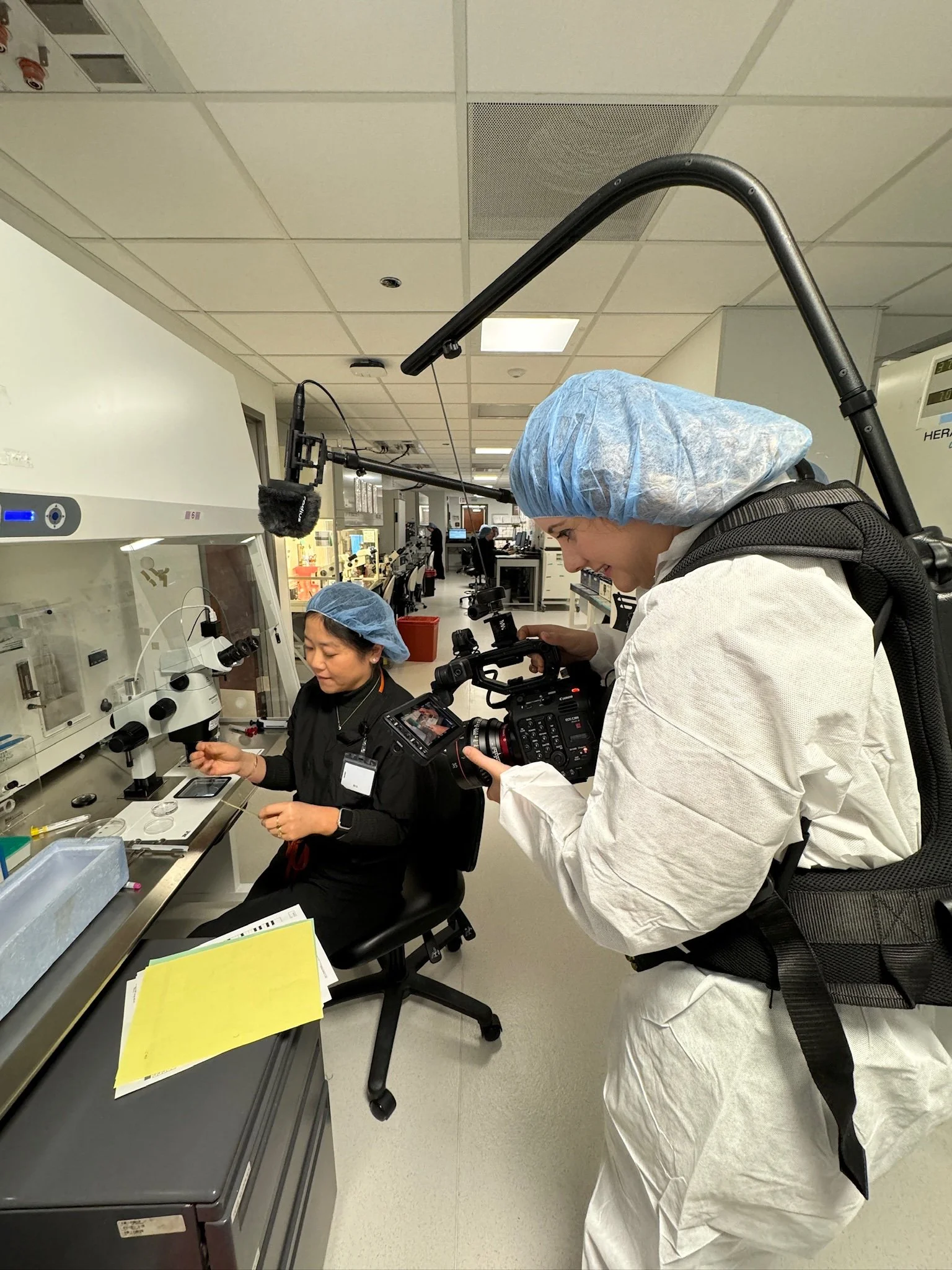 A person wearing a blue hair cover and a white lab coat is filming a woman working with laboratory equipment in a scientific or medical setting.